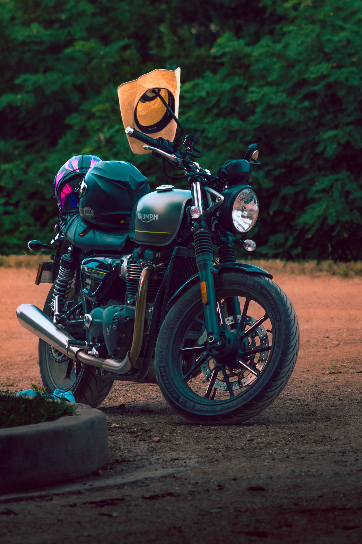 Dark green cafe racer motorcycle parked on dirt ground at dusk with moody lighting and forest background.
