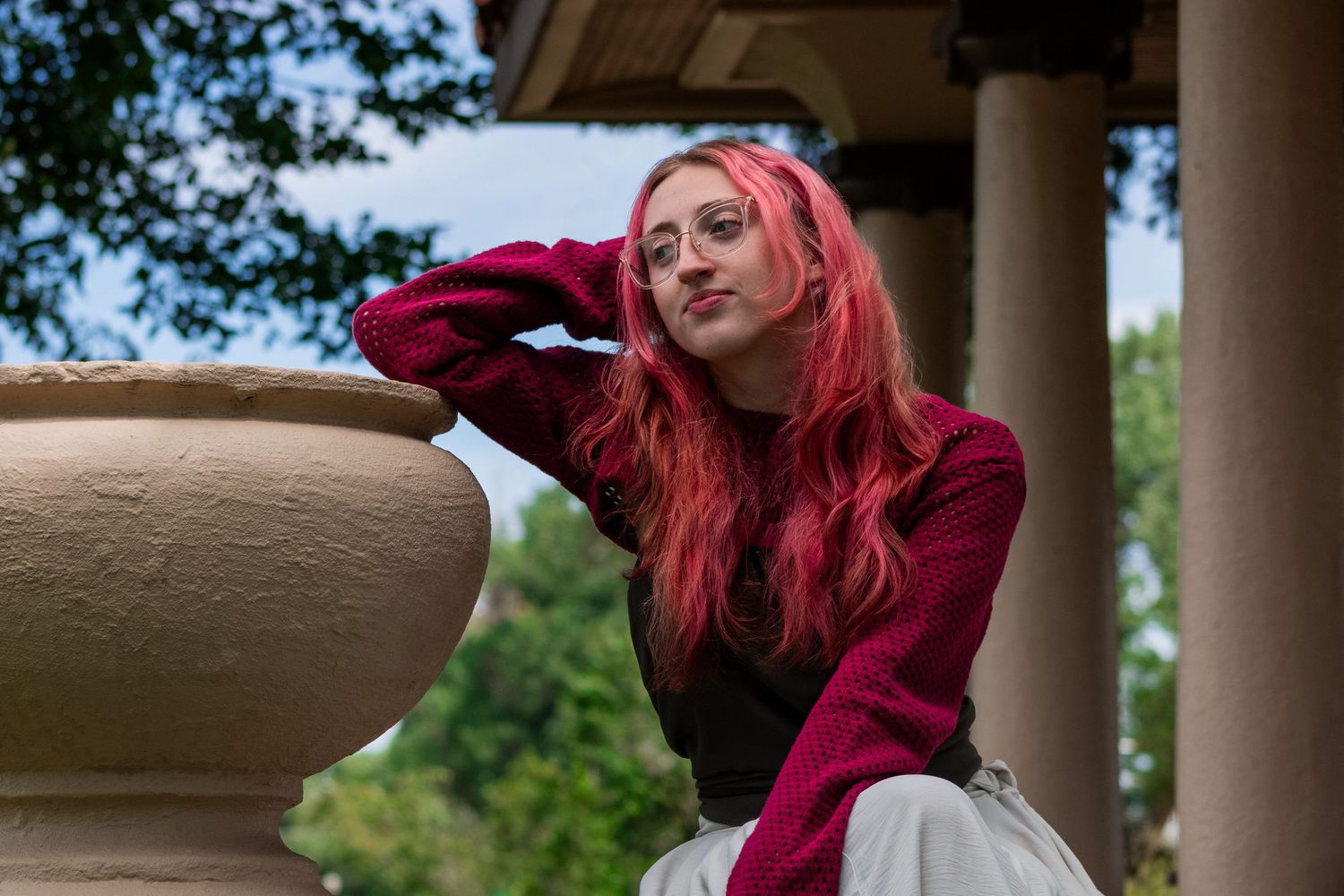 Figure in red sweater poses elegantly by stone pillar in outdoor setting.