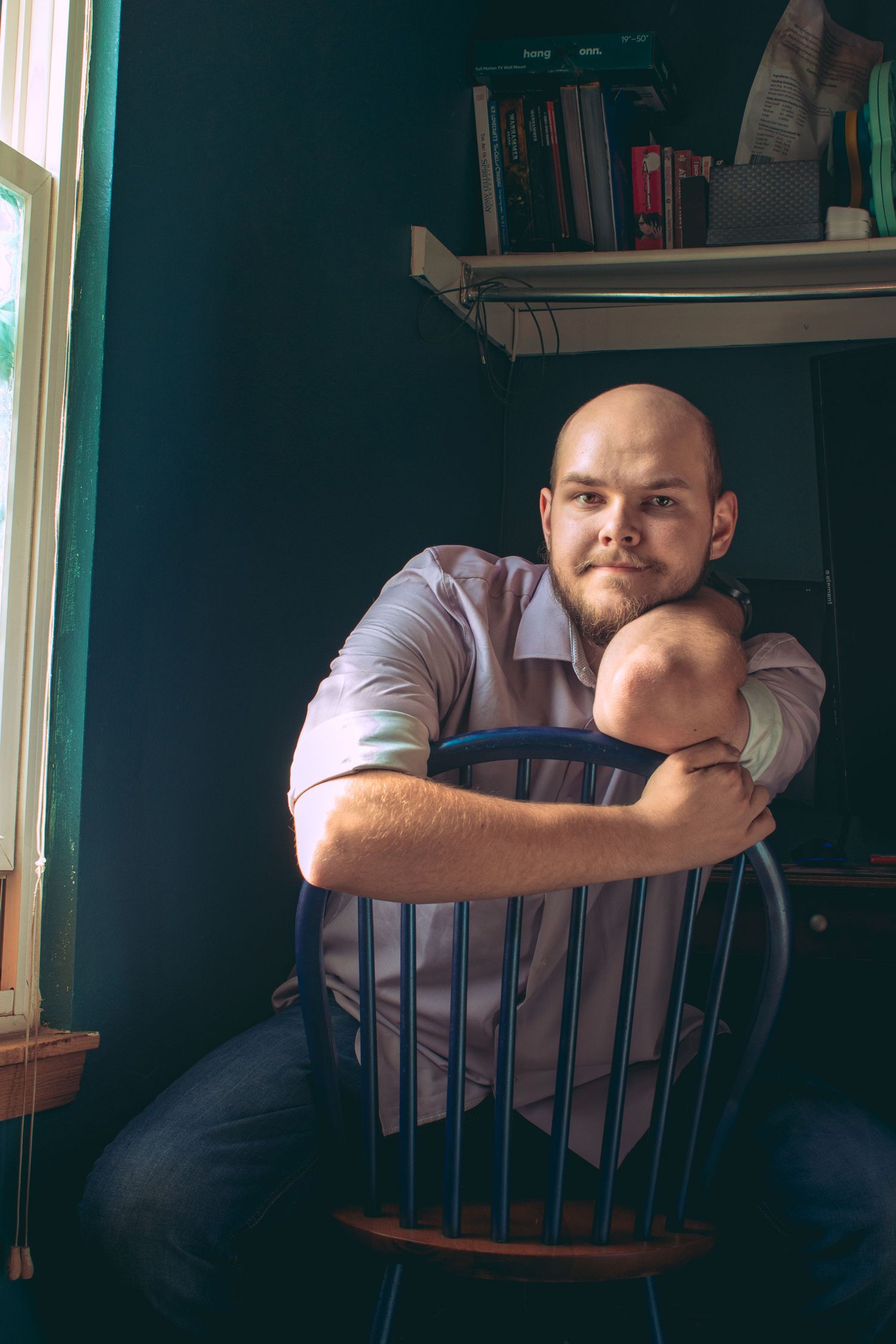 Low-lit indoor portrait showing someone seated in a wooden chair near bookshelves.
