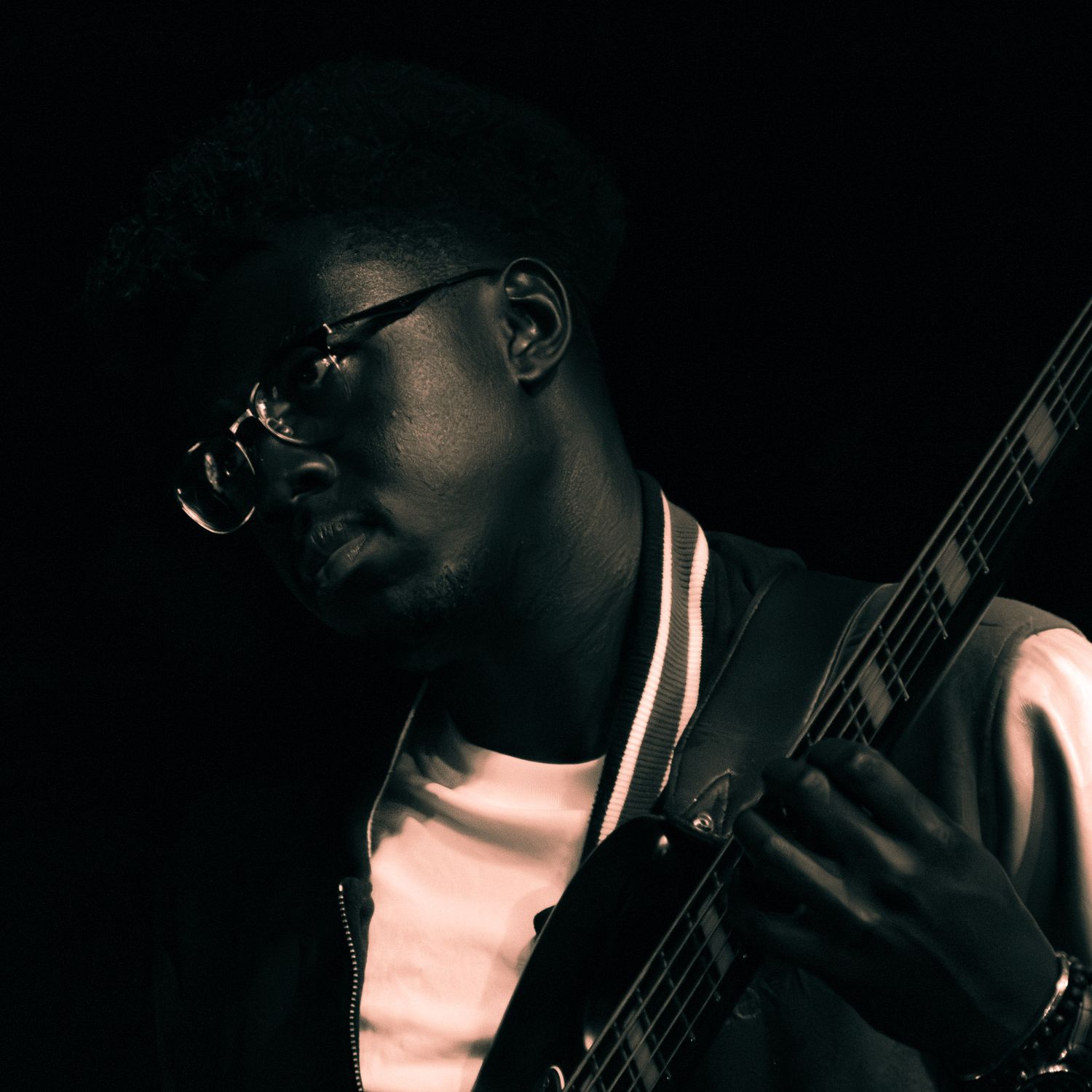 A young man with glasses plays an electric guitar in a dark studio setting, wearing a varsity jacket.