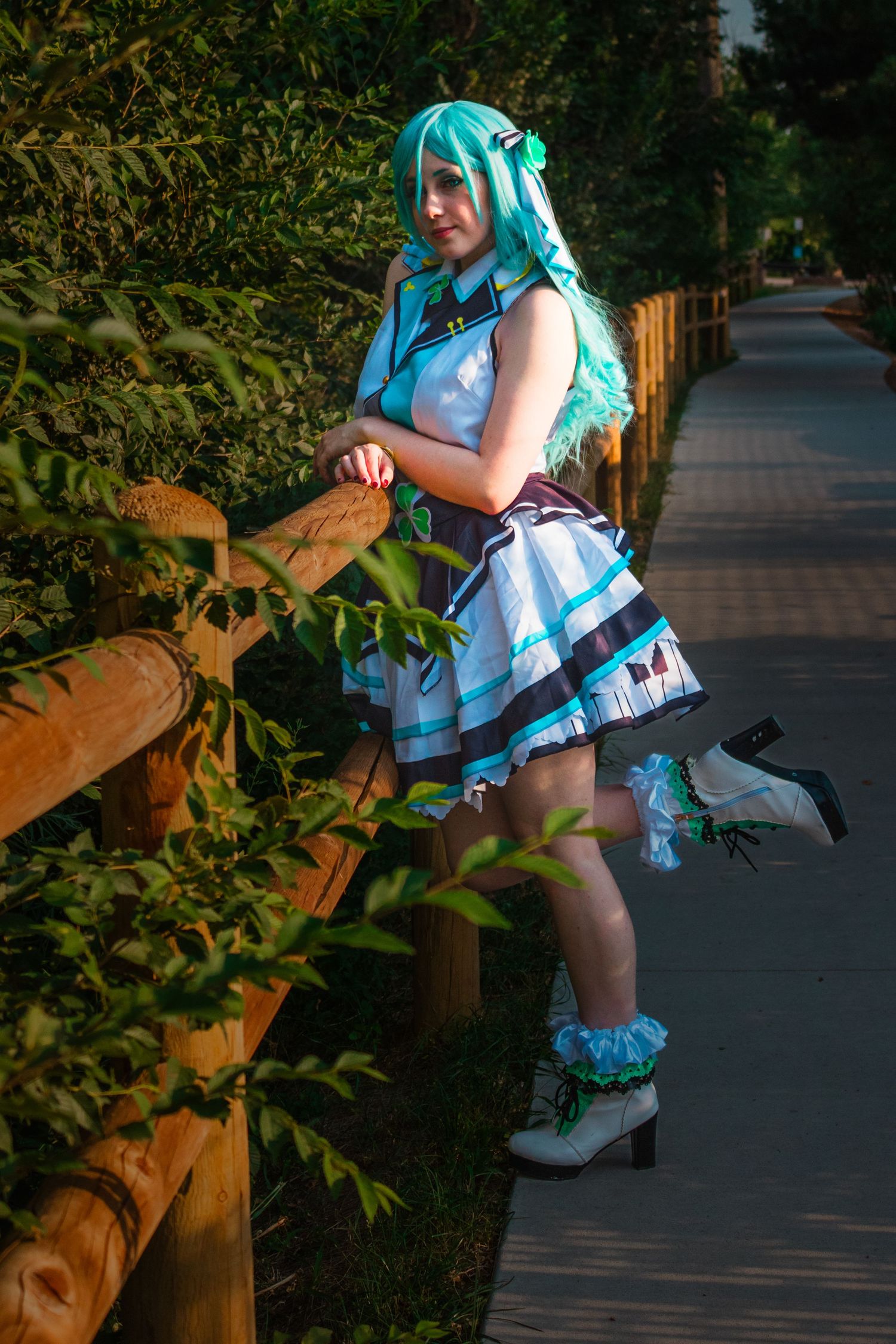 Cosplay model in turquoise wig and matching dress poses playfully along garden path with wooden railing.
