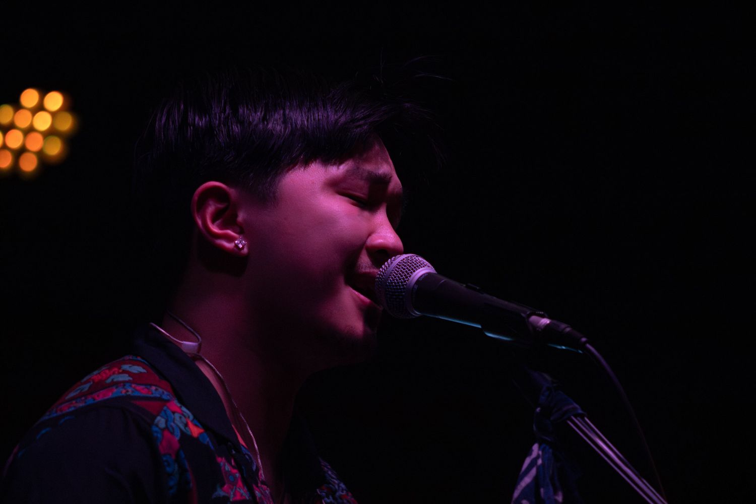 Young musician singing into a microphone on a dark stage with warm bokeh lights in the background.