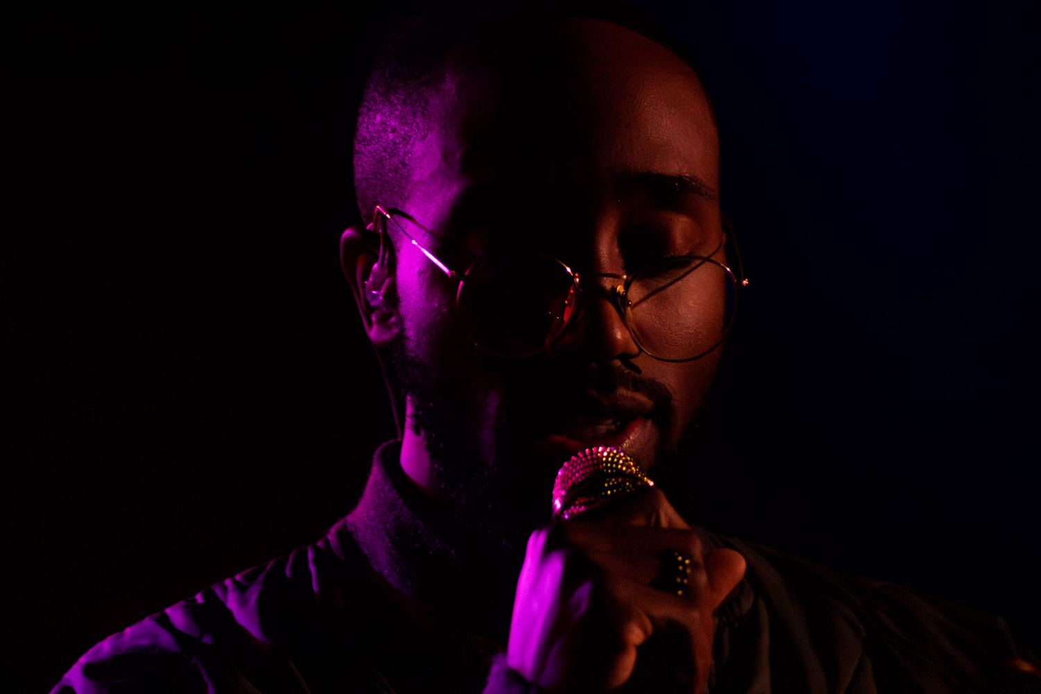 Man with glasses singing into microphone under dramatic purple and pink stage lighting in dark setting.