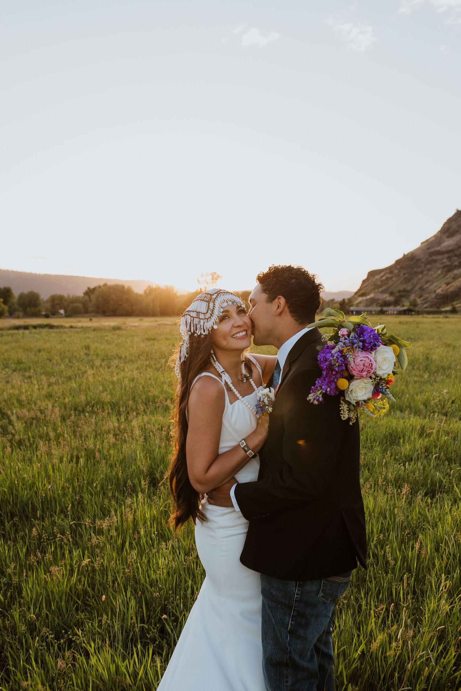 A romantic wedding couple shares intimate moments at sunset in a scenic meadow with mountains in the background.