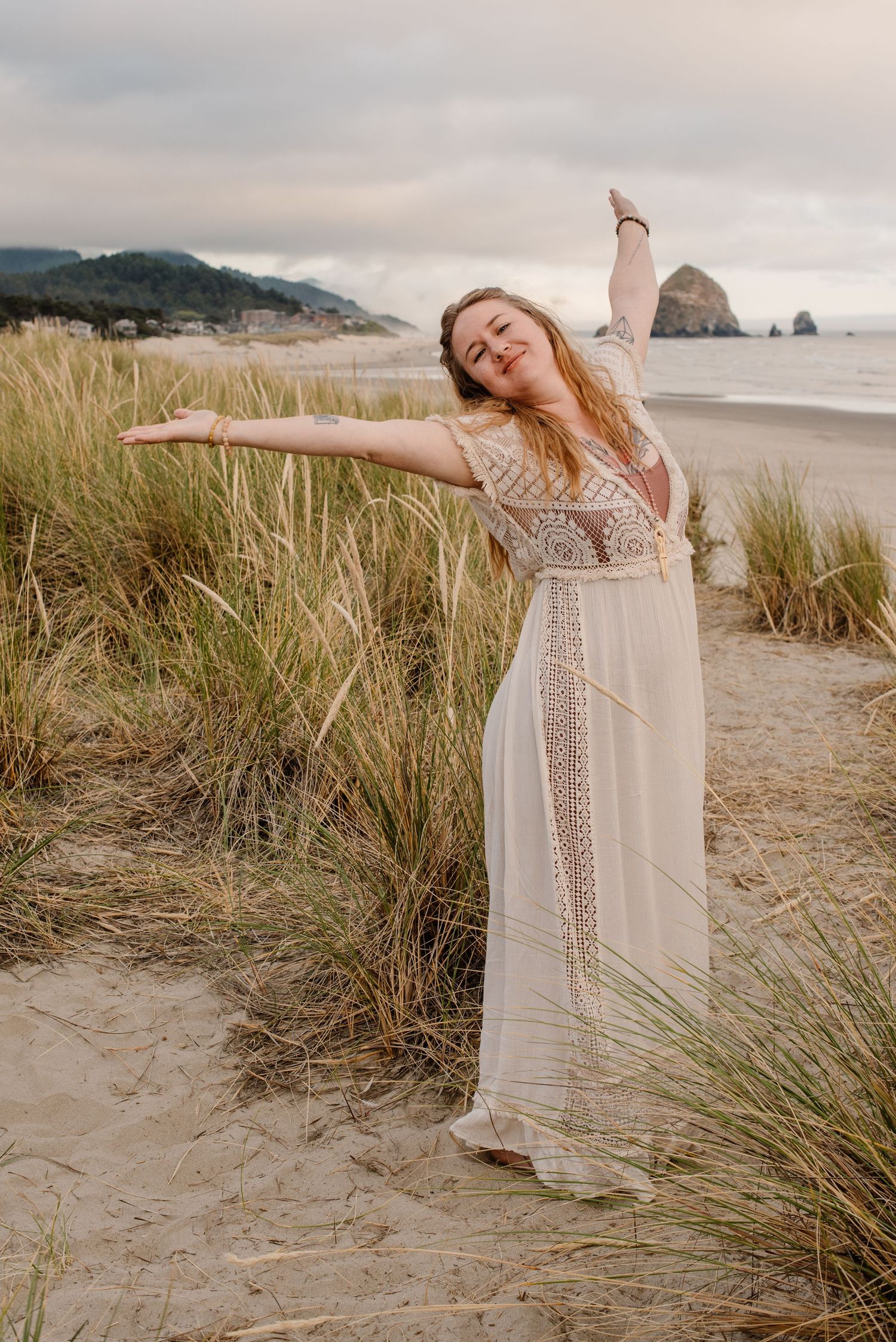 Woman in flowy white dress poses expressively on beach with coastal dunes and grasses at sunset.