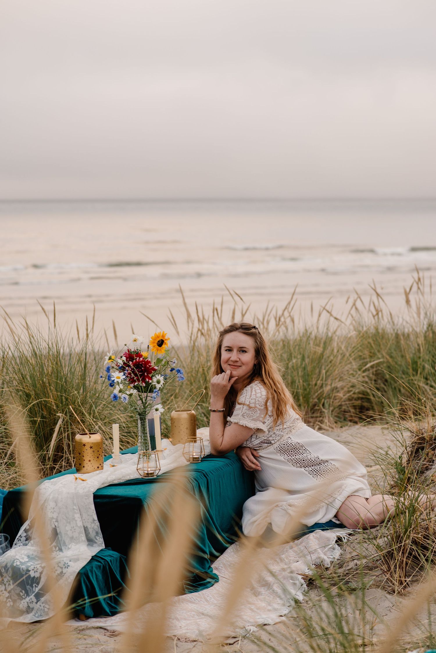 A romantic beach picnic setup with emerald linens, wildflowers and ocean views at sunset through the beach grass.