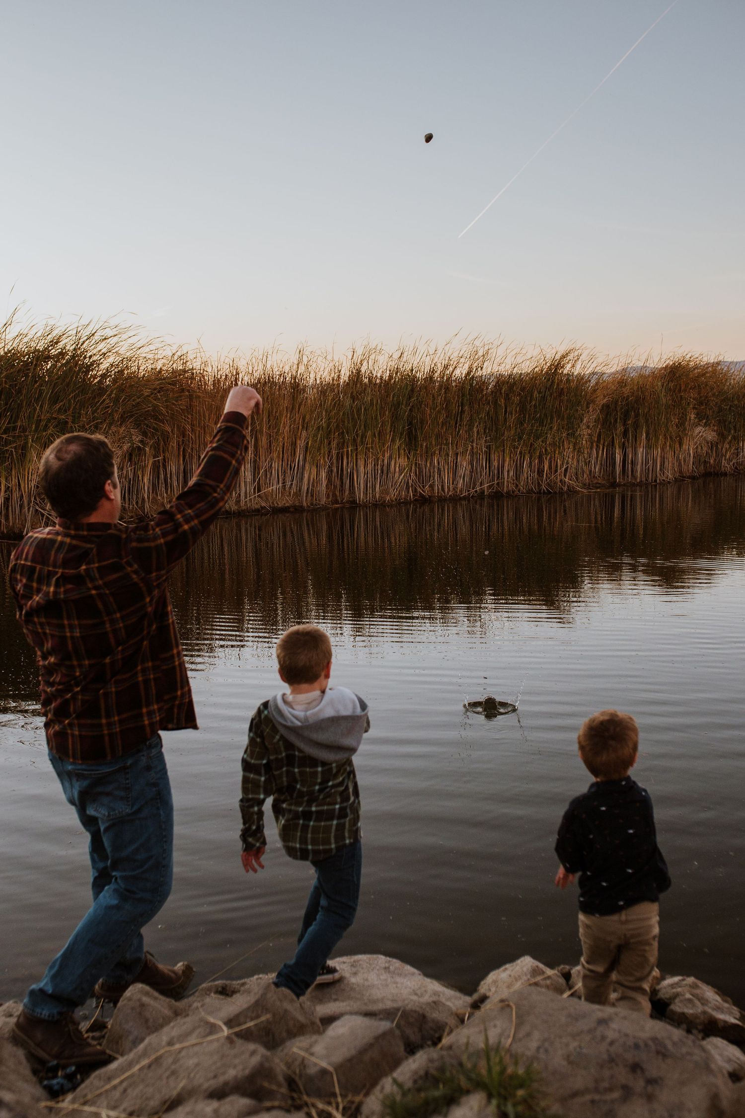 Three people stand on rocks by a lake at sunset while tossing something into the calm water with tall reeds in background.
