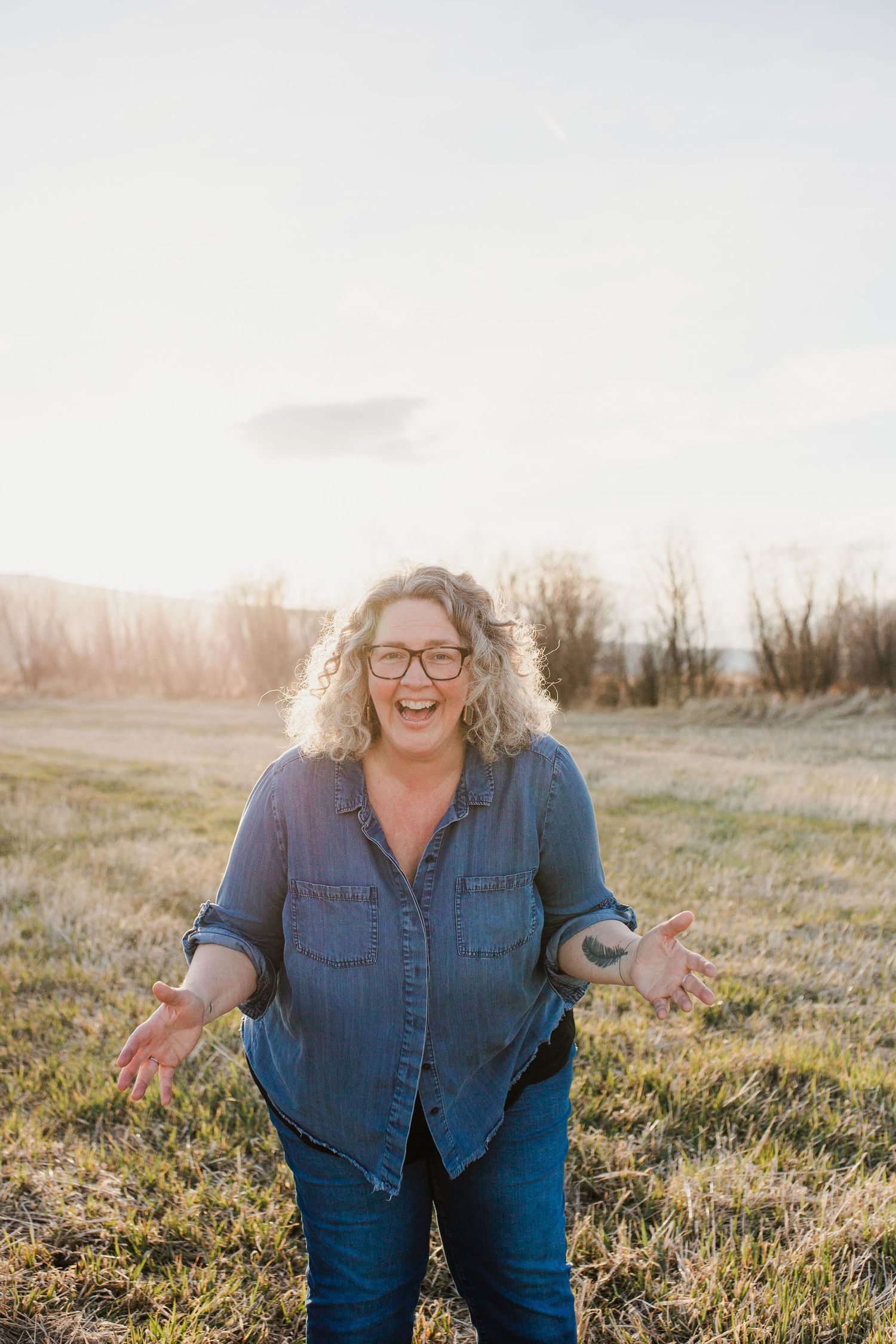 A person in a denim shirt stands in a sunlit field with grass during a warm golden hour.