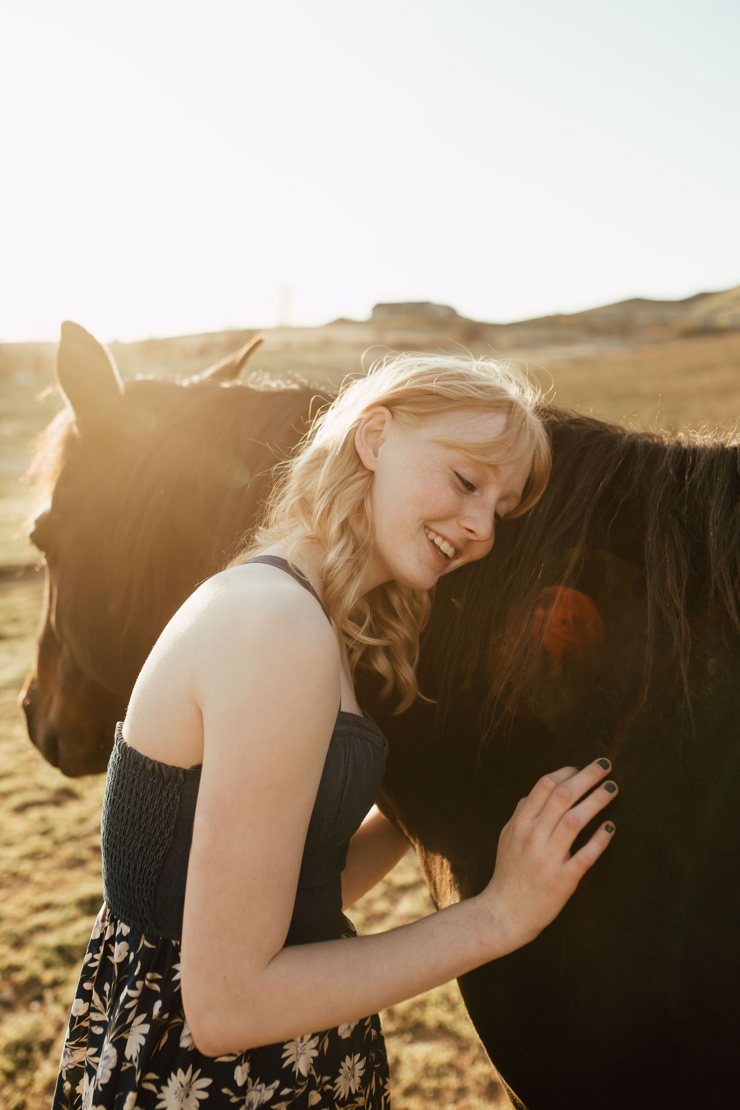 A person in a black and white dress caresses a dark horse in warm golden sunlight.