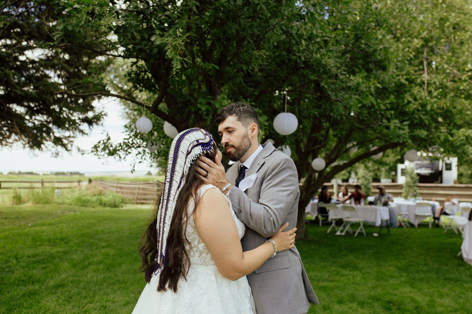 A couple shares a tender moment during their outdoor wedding reception on a green lawn with white chairs and string lights.
