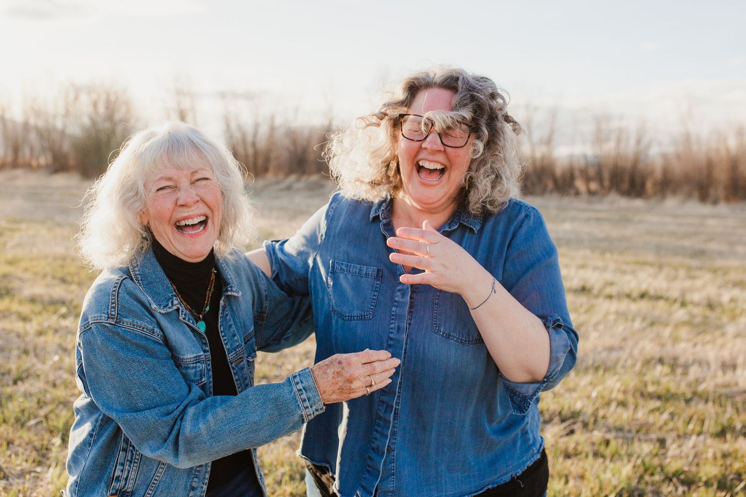 Mother and daughter in denim jackets share joyful laughter while together outdoors in golden sunlight.