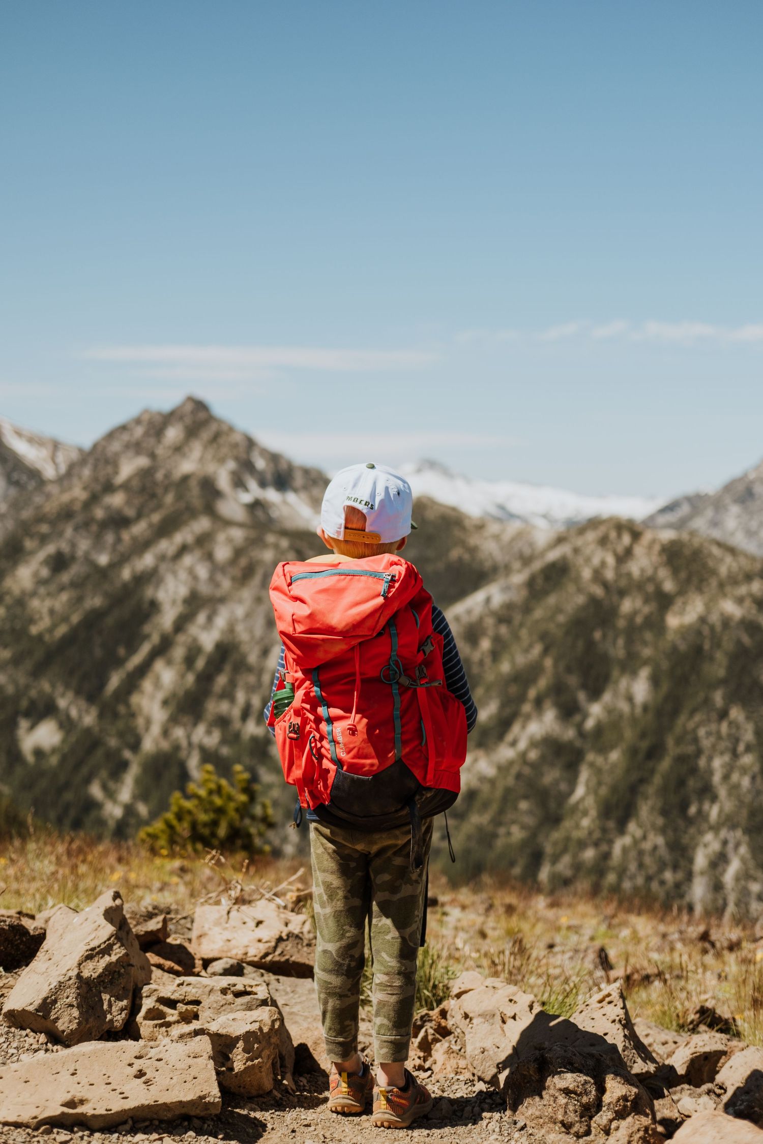 Sequence of a hiker with bright red backpack and tan boots standing on rocky mountain terrain against blue skies.