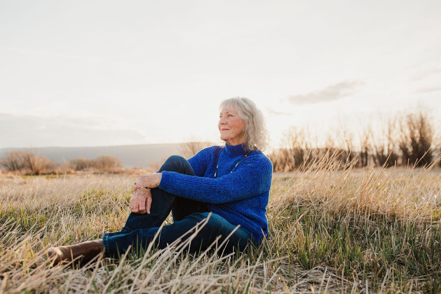 Person wearing blue sweater relaxes in golden grass field at sunset with mountains in background.