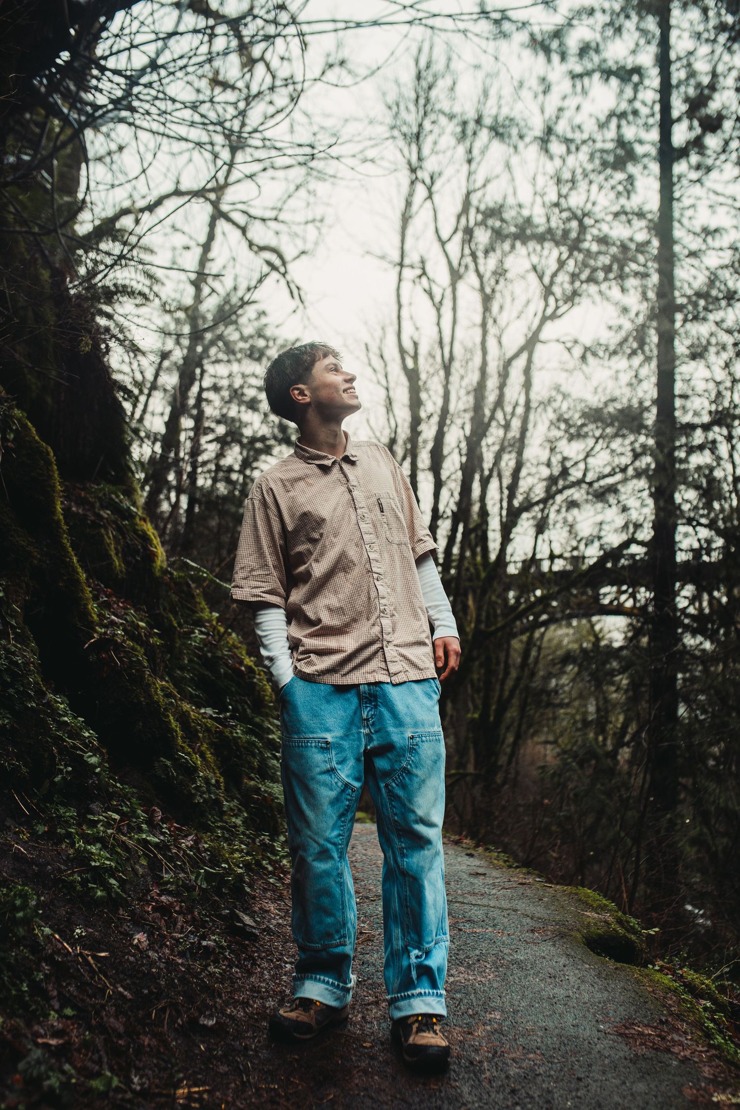 A person in blue jeans and beige sweater stands on a mossy forest path looking up at the trees on an overcast day.