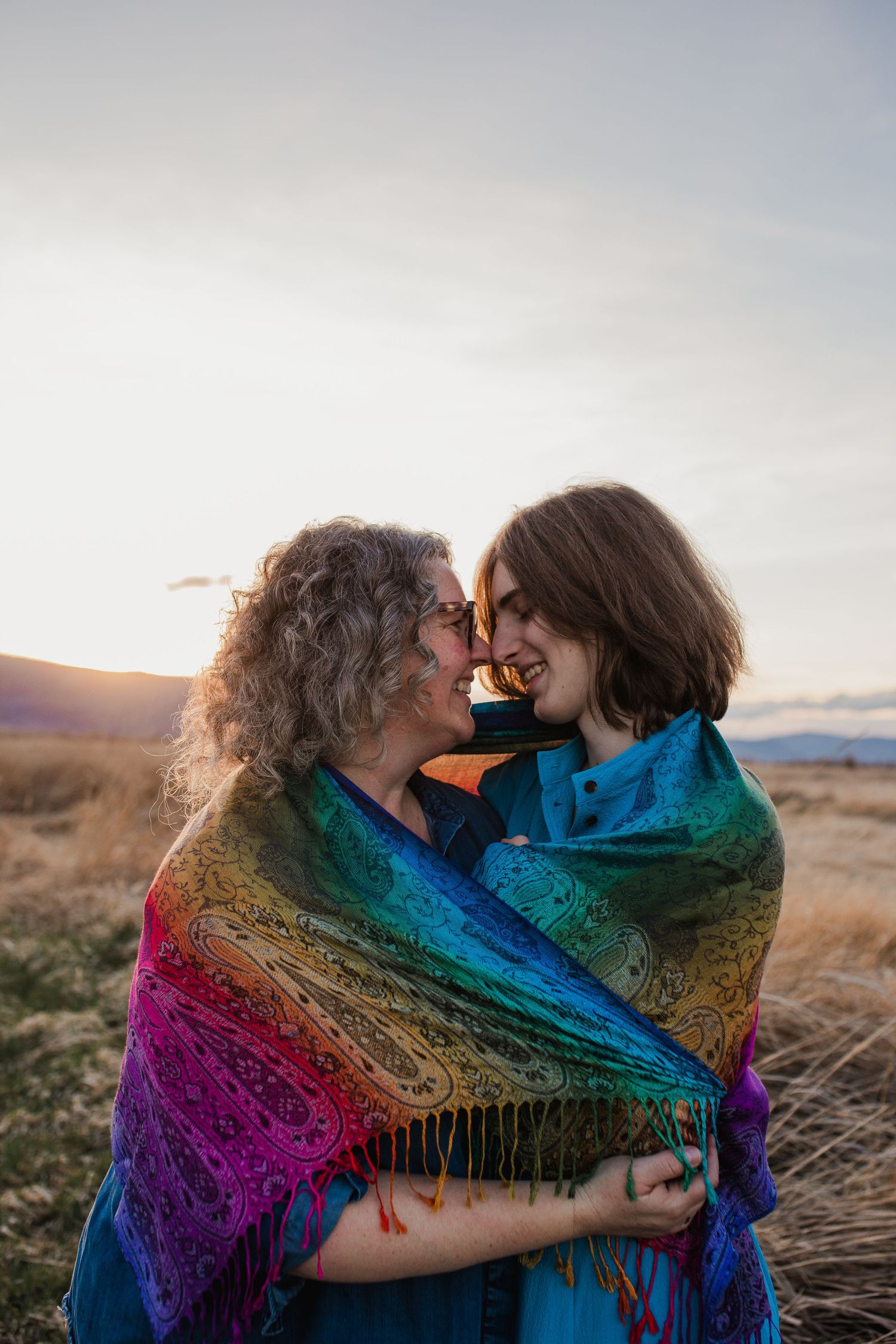 Two figures wrapped in a multicolored rainbow shawl embrace on a grassy hillside at sunset.