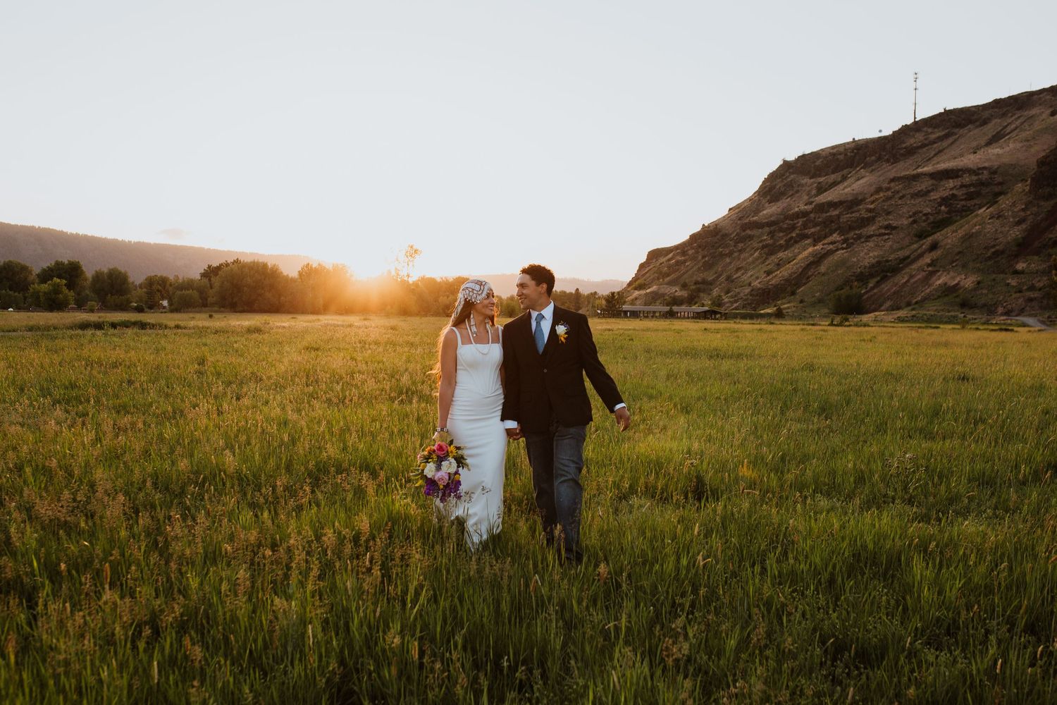 A wedding couple walks through a golden field at sunset with rolling hills in the background.