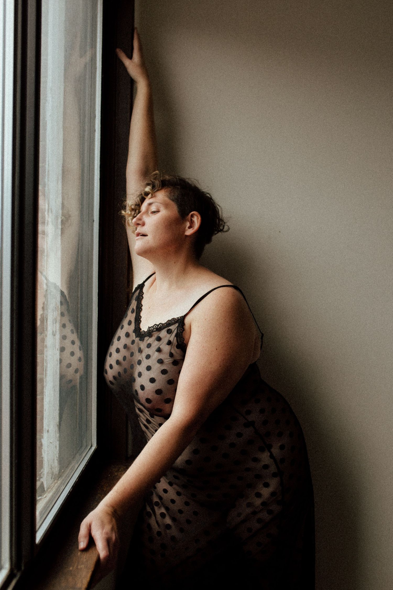 A figure in a polka dot dress poses contemplatively by a window in a series of moody portrait shots.