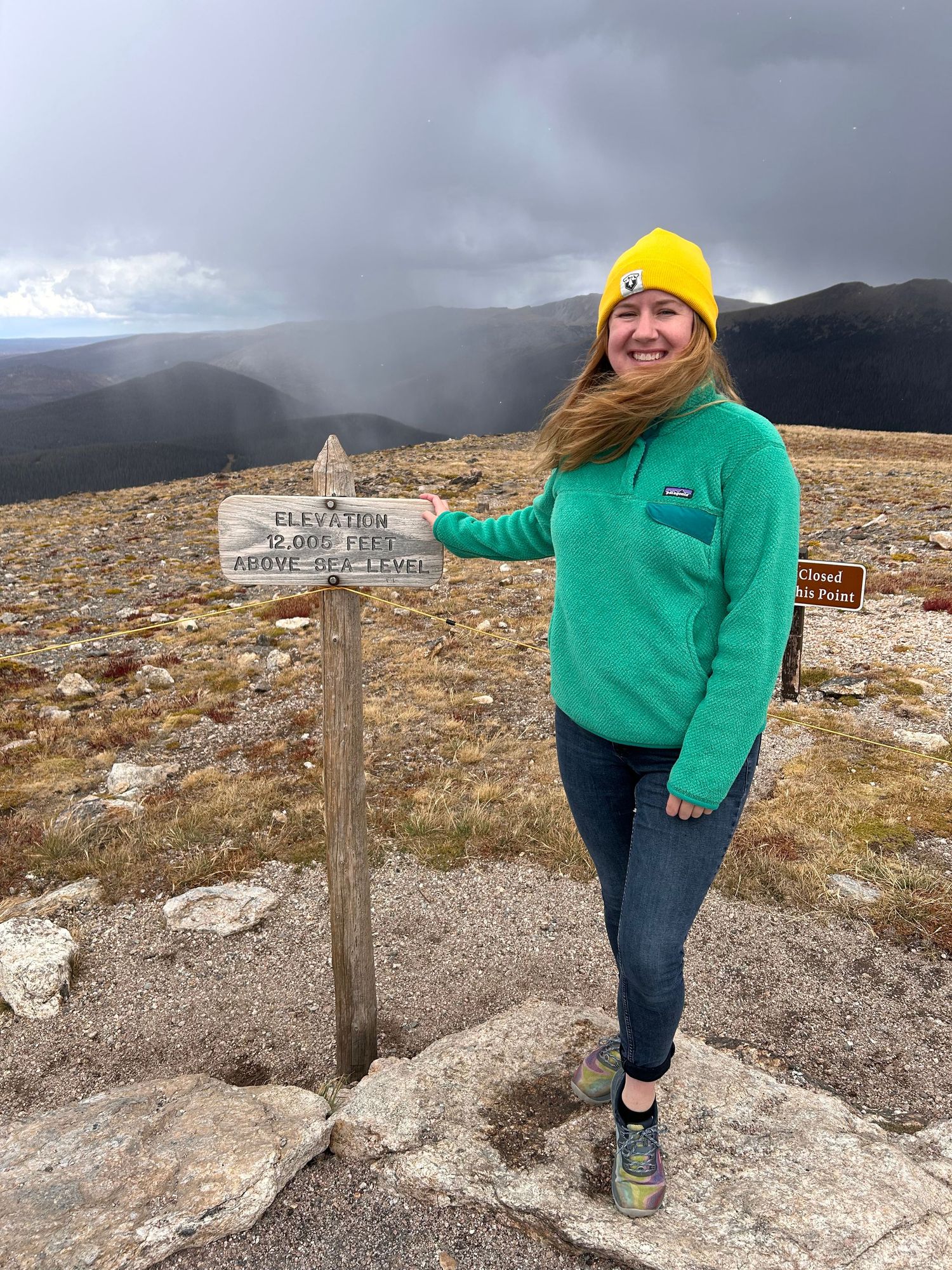 A hiker in a bright yellow beanie and green jacket poses near a mountain trail marker sign against a cloudy sky.