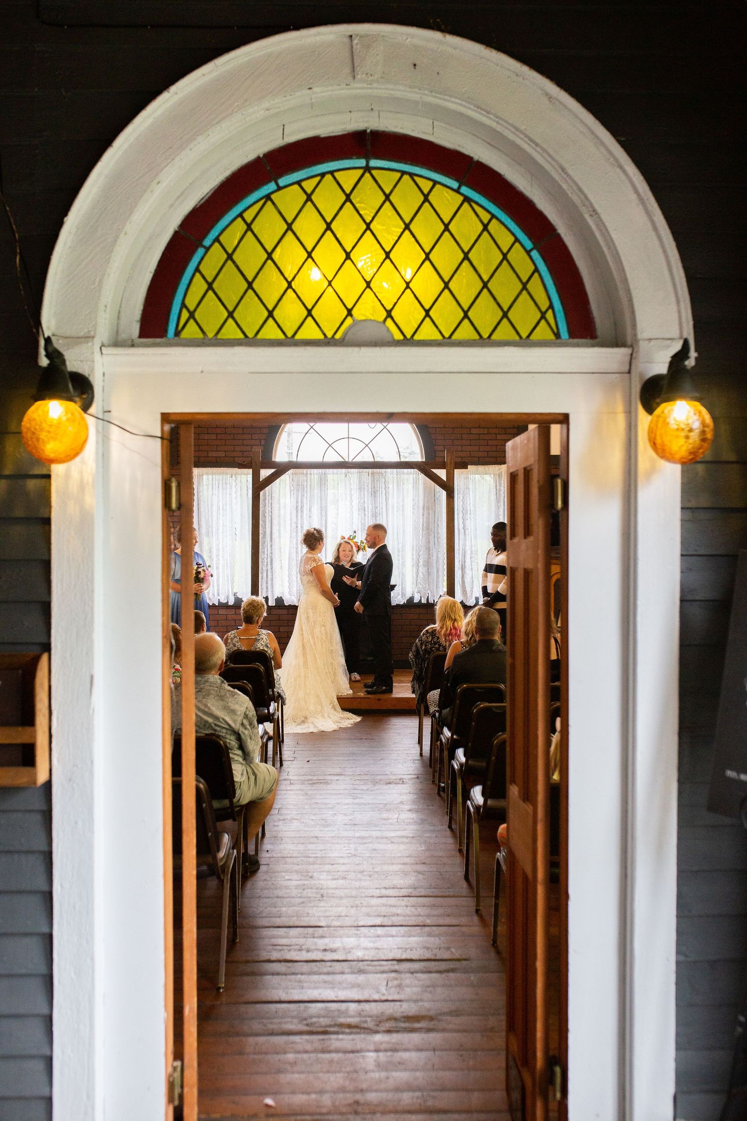 Long church aisle with wooden pews and yellow arched stained glass window at the end of sunlit corridor.