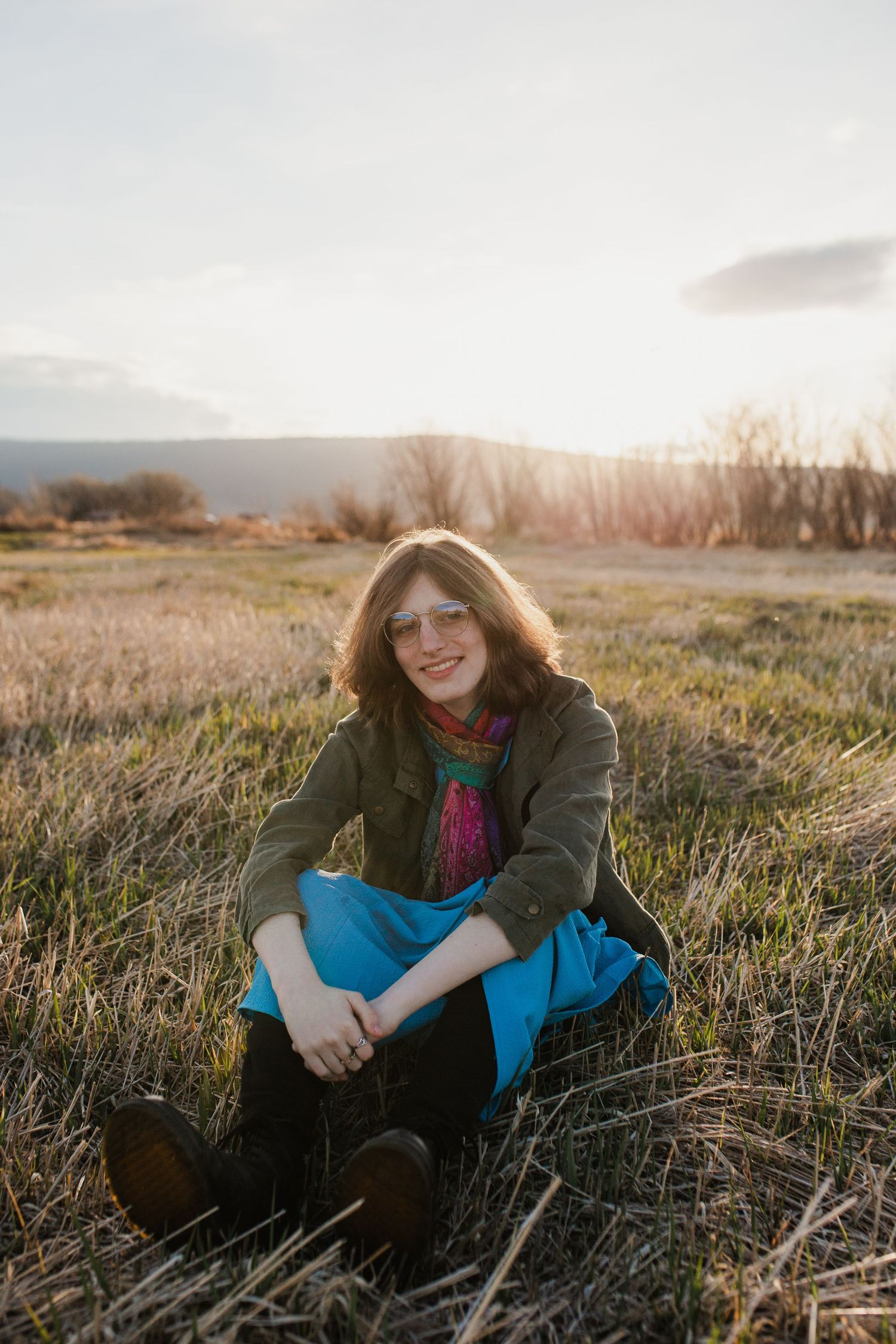 Person in olive jacket and colorful scarf sits in golden field at sunset with soft natural lighting and warm autumn tones.