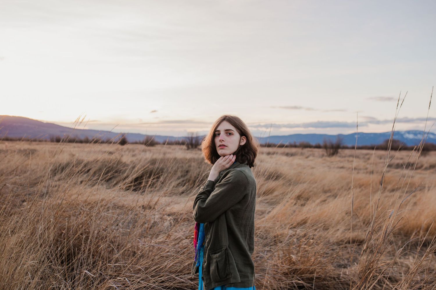 Person with long hair wearing an olive jacket walks through tall golden grass in a rural mountainous landscape at sunset.