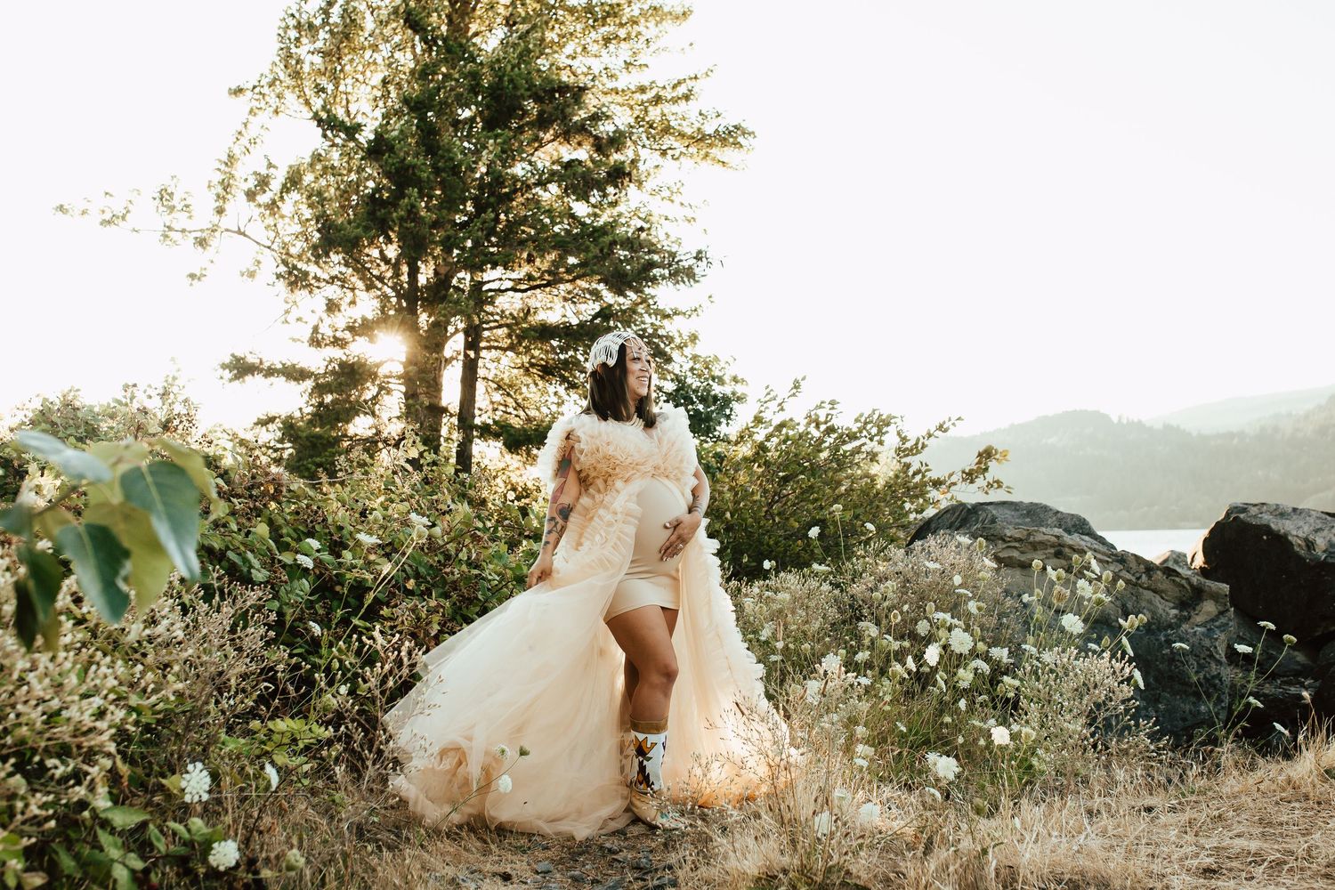 A pregnant woman in a flowing white dress stands on a hilltop with mountain and lake views during golden hour sunset.