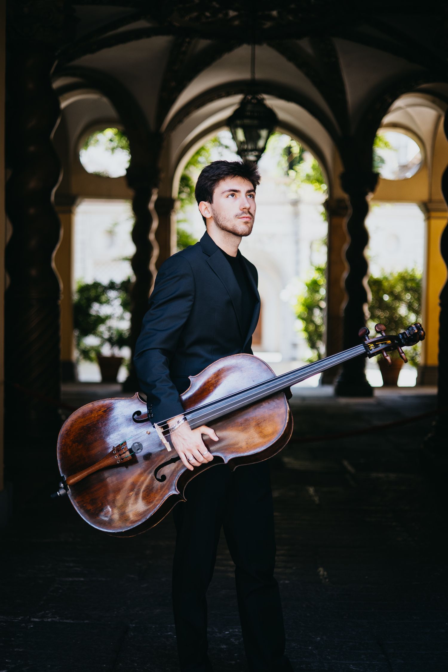 Young male cellist in black jacket holds a cello under arched stone columns in a sunlit outdoor classical setting.