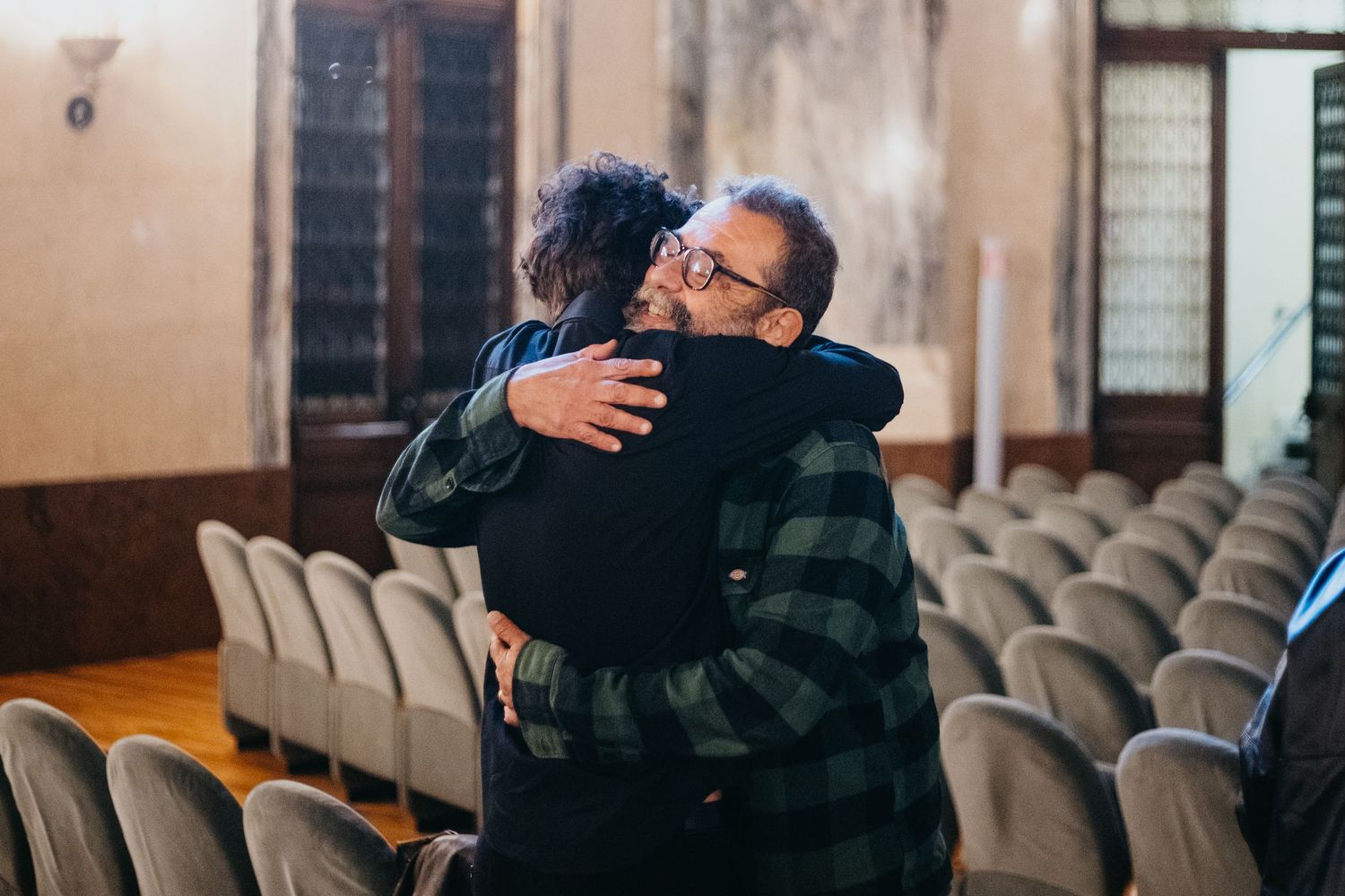 Two people share a warm embrace in a hall with rows of empty chairs at a formal event.