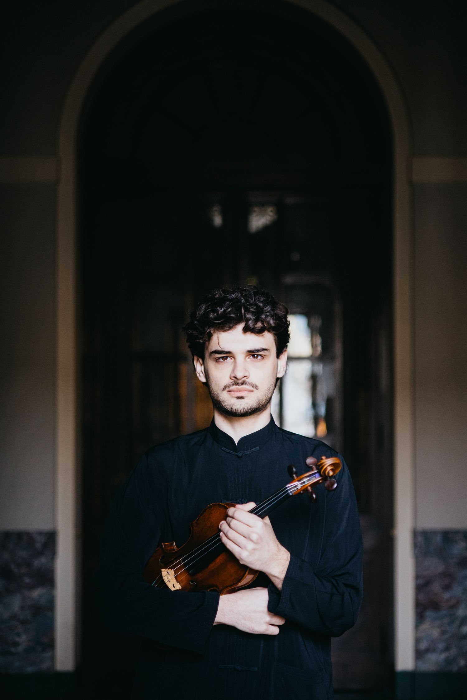 Young male violinist in black outfit holds violin while standing in arched doorway corridor.