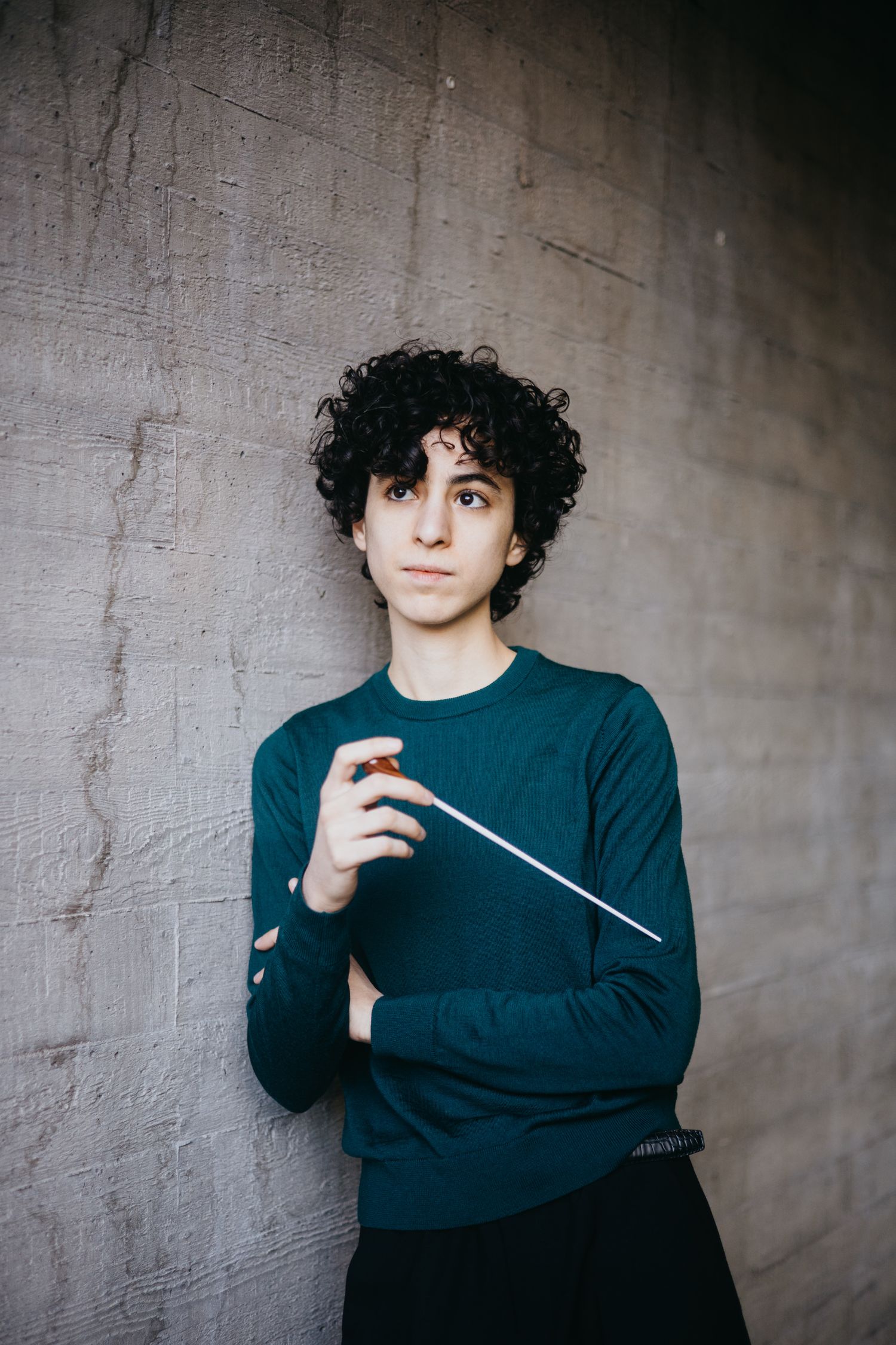 Curly-haired conductor holding baton against gray concrete wall outdoors.