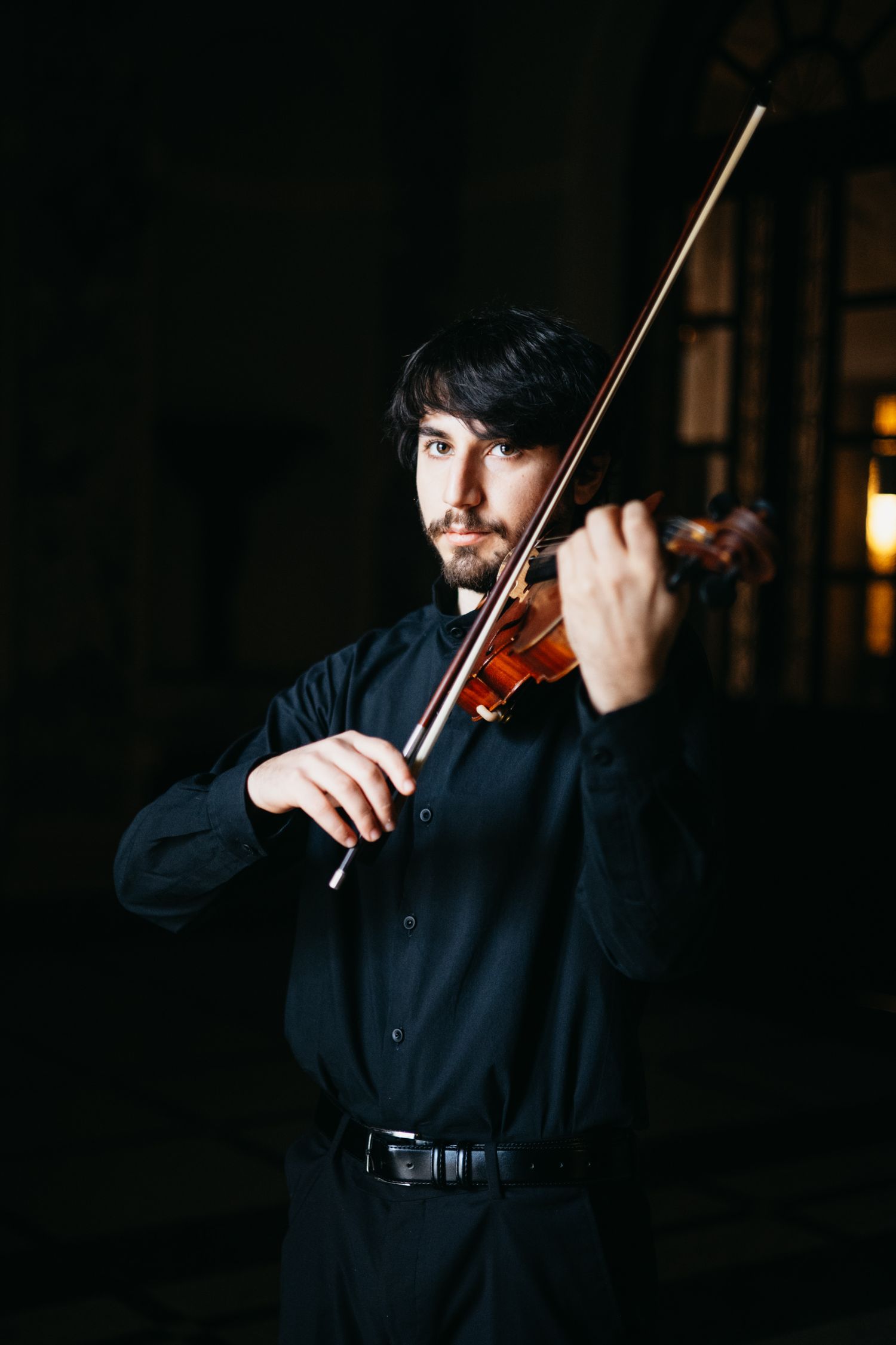 Young male musician playing violin passionately in black outfit against dark background.