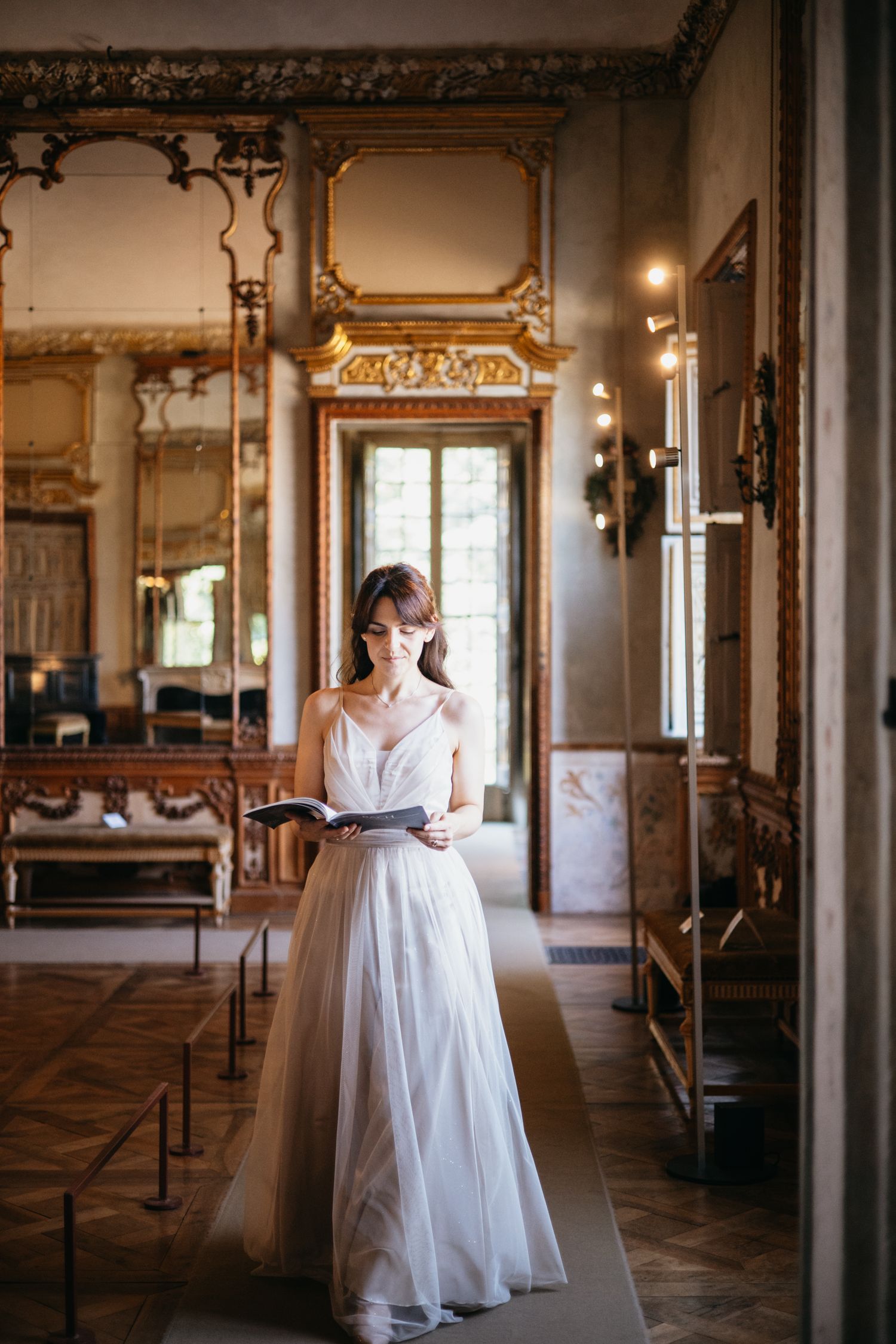 Bride in white gown reads a book in an ornate baroque hall with gilded mirrors and marble decor.
