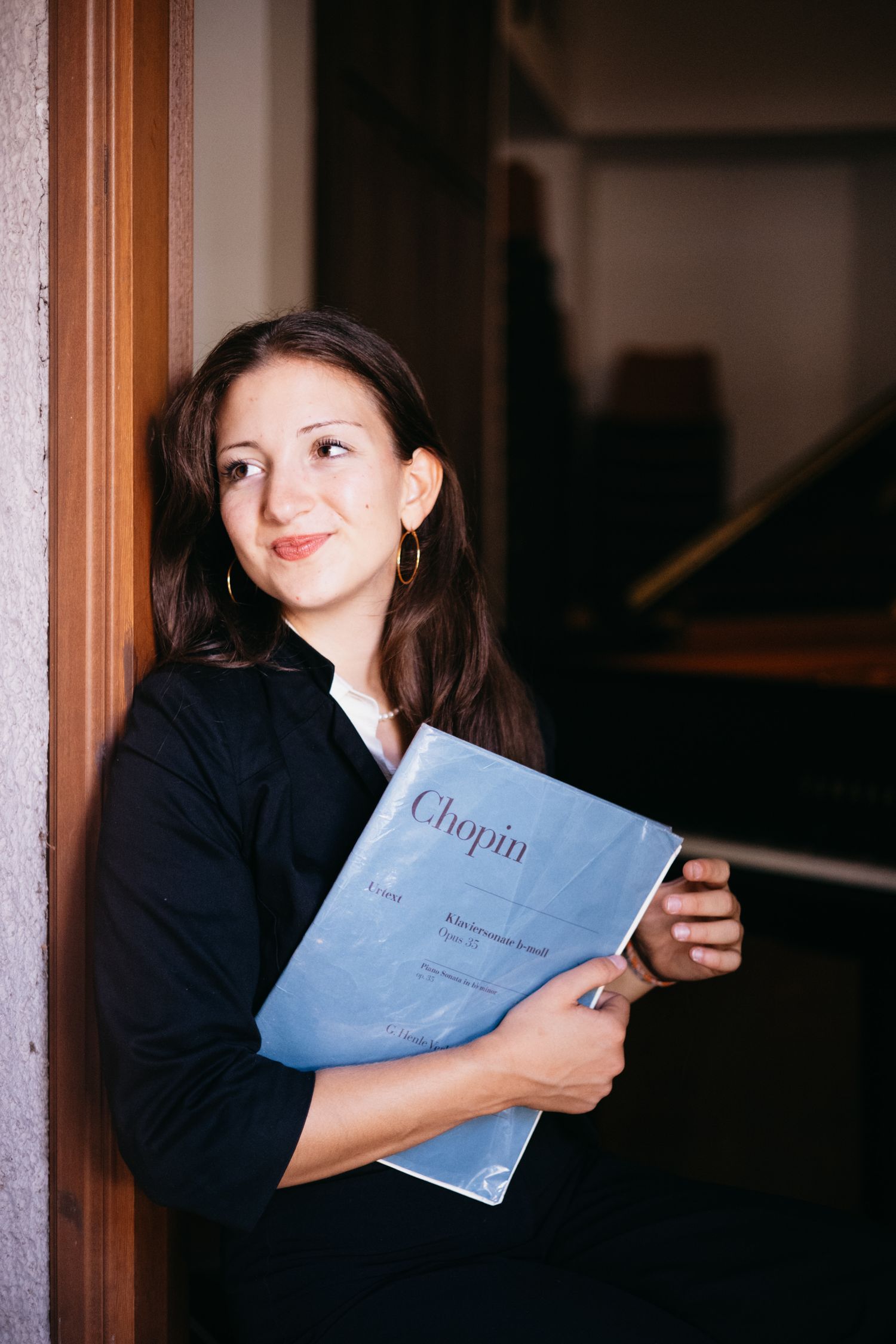 Young woman smiling and holding a Chopin sheet music book, leaning against a wall near a piano.