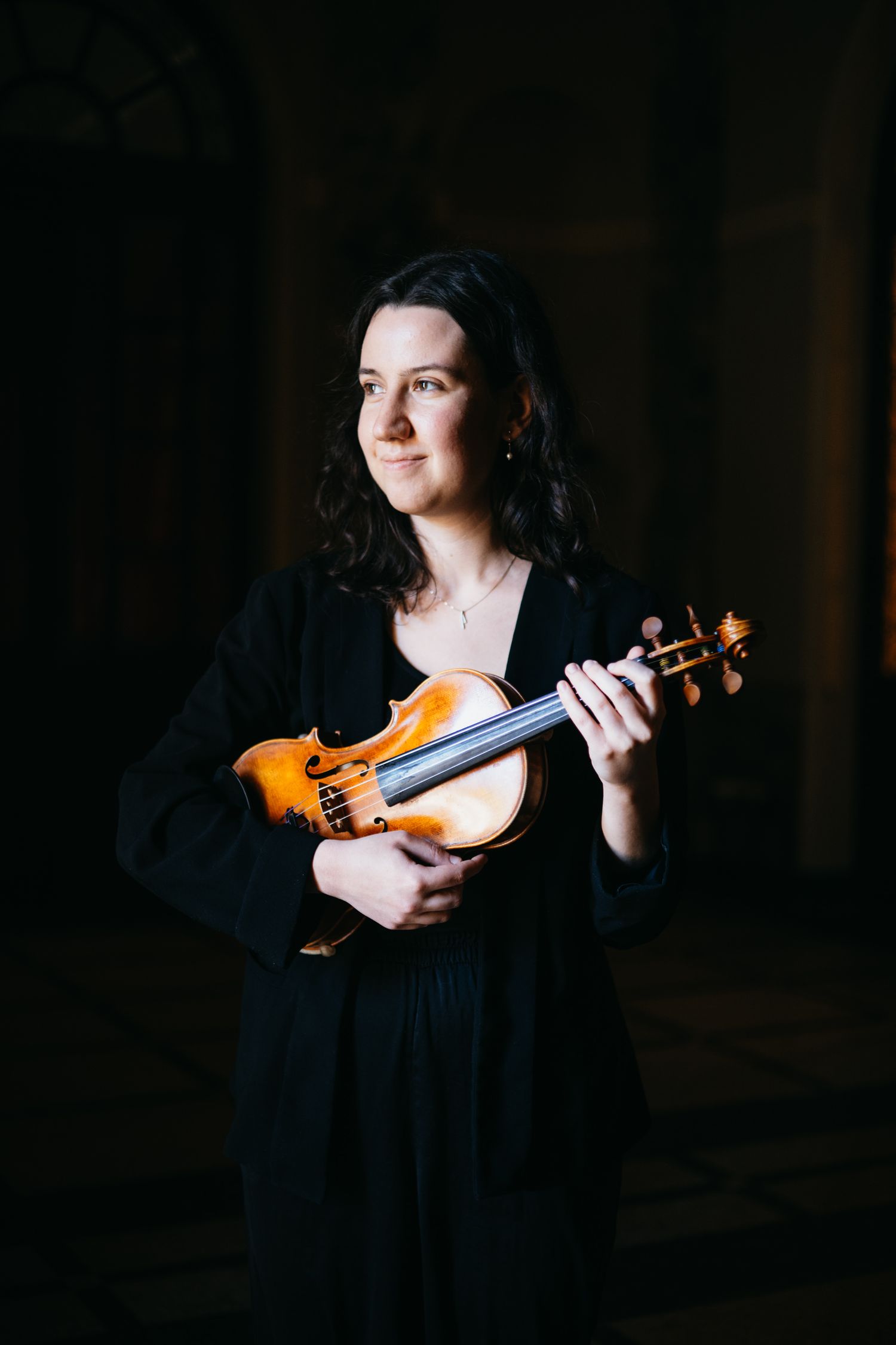 Woman in black dress holding a violin, posing elegantly in a dark, dramatic setting.