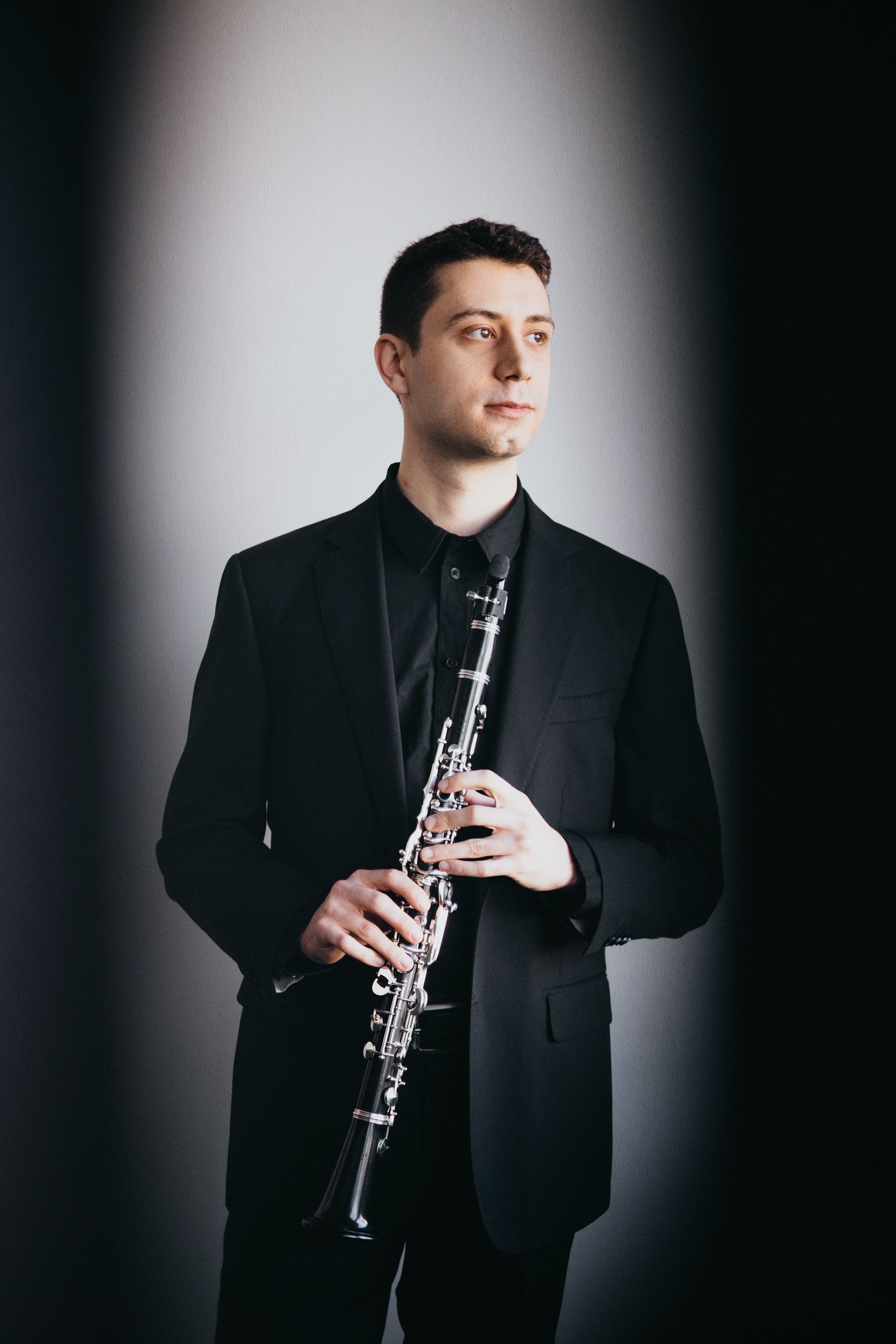 A young male musician in a black suit holds a clarinet while posing against a studio backdrop.