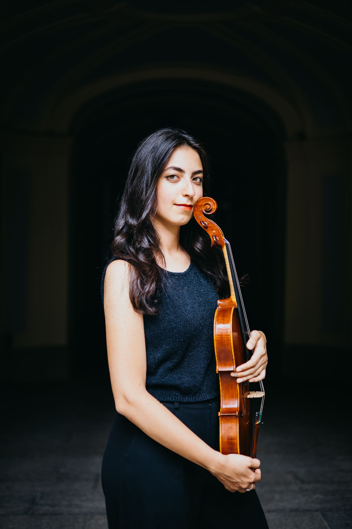 Young woman in black sleeveless dress holds violin upright against dark arched backdrop.