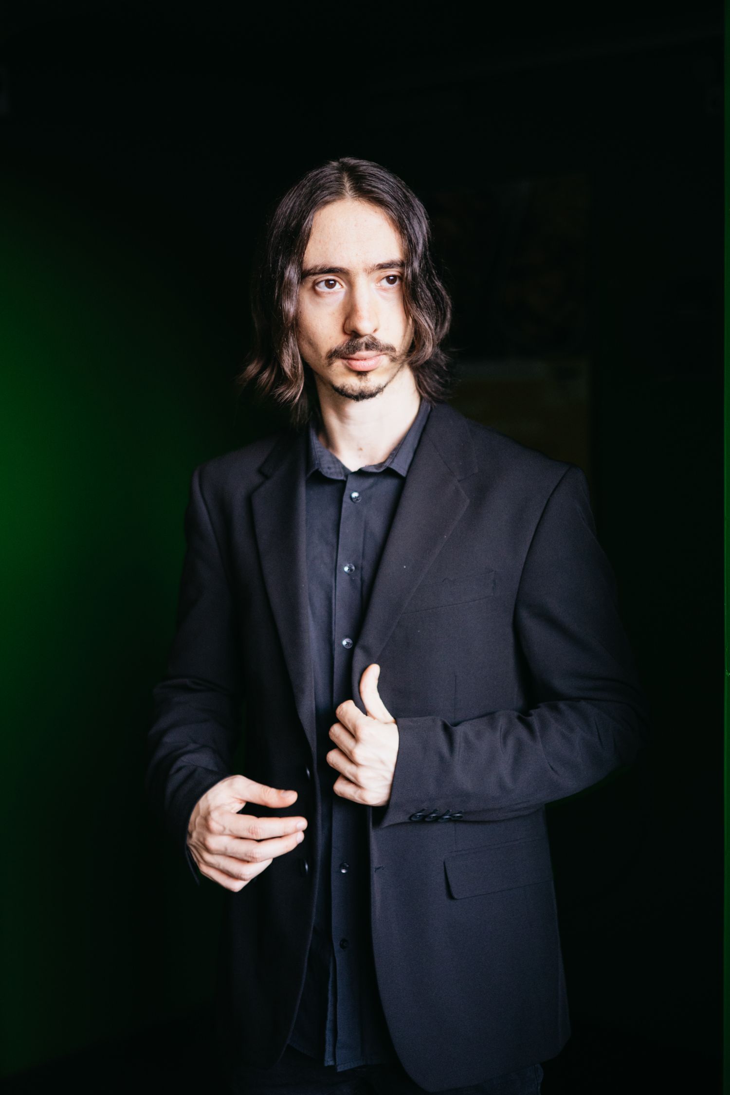 Long-haired male pianist in black blazer posing confidently against a dark green background.