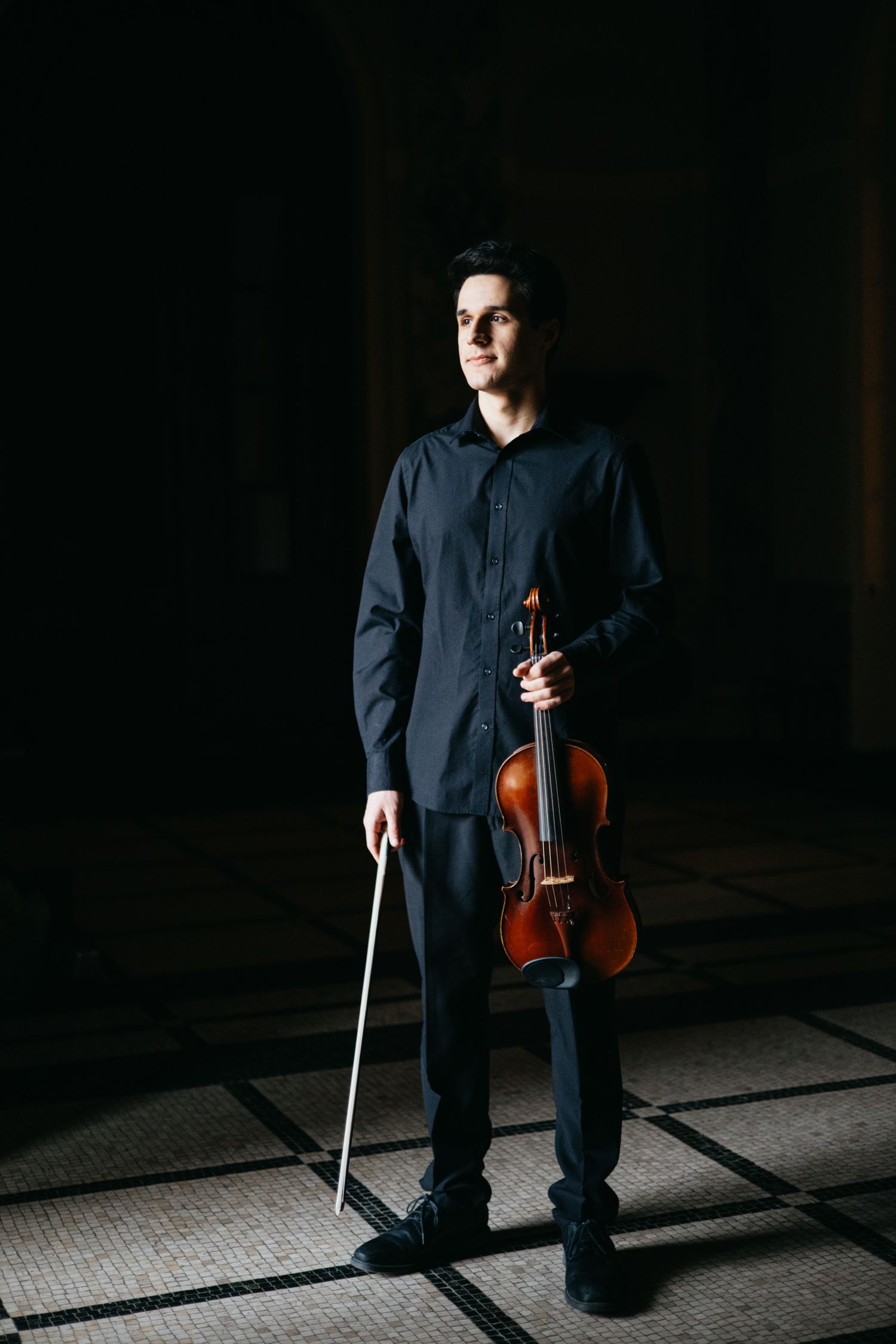 Male violinist stands holding violin and bow in full-length dark atmospheric portrait.