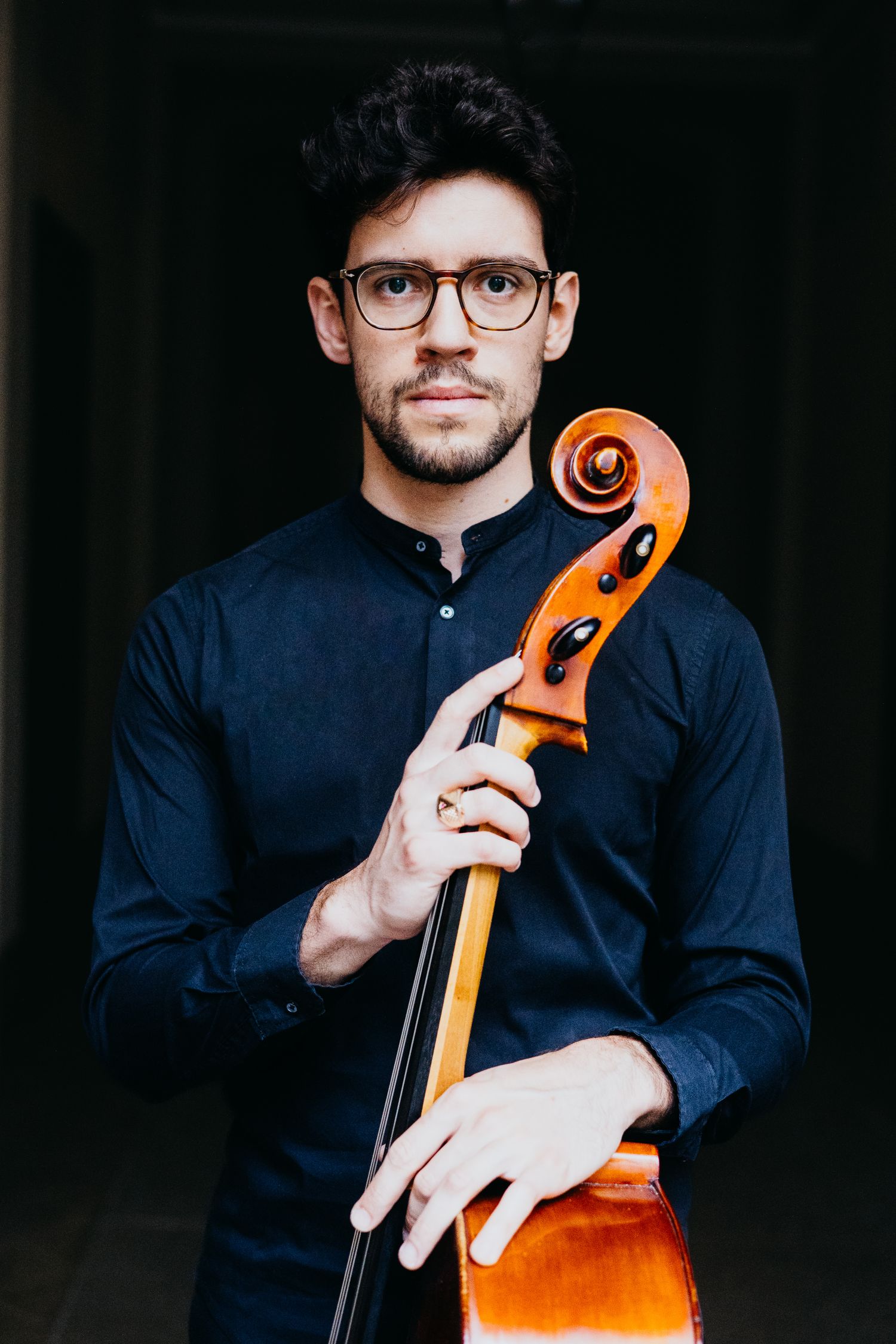 Violoncellista con gli occhiali e camicia scura, mentre tiene il manico del violoncello su uno sfondo scuro