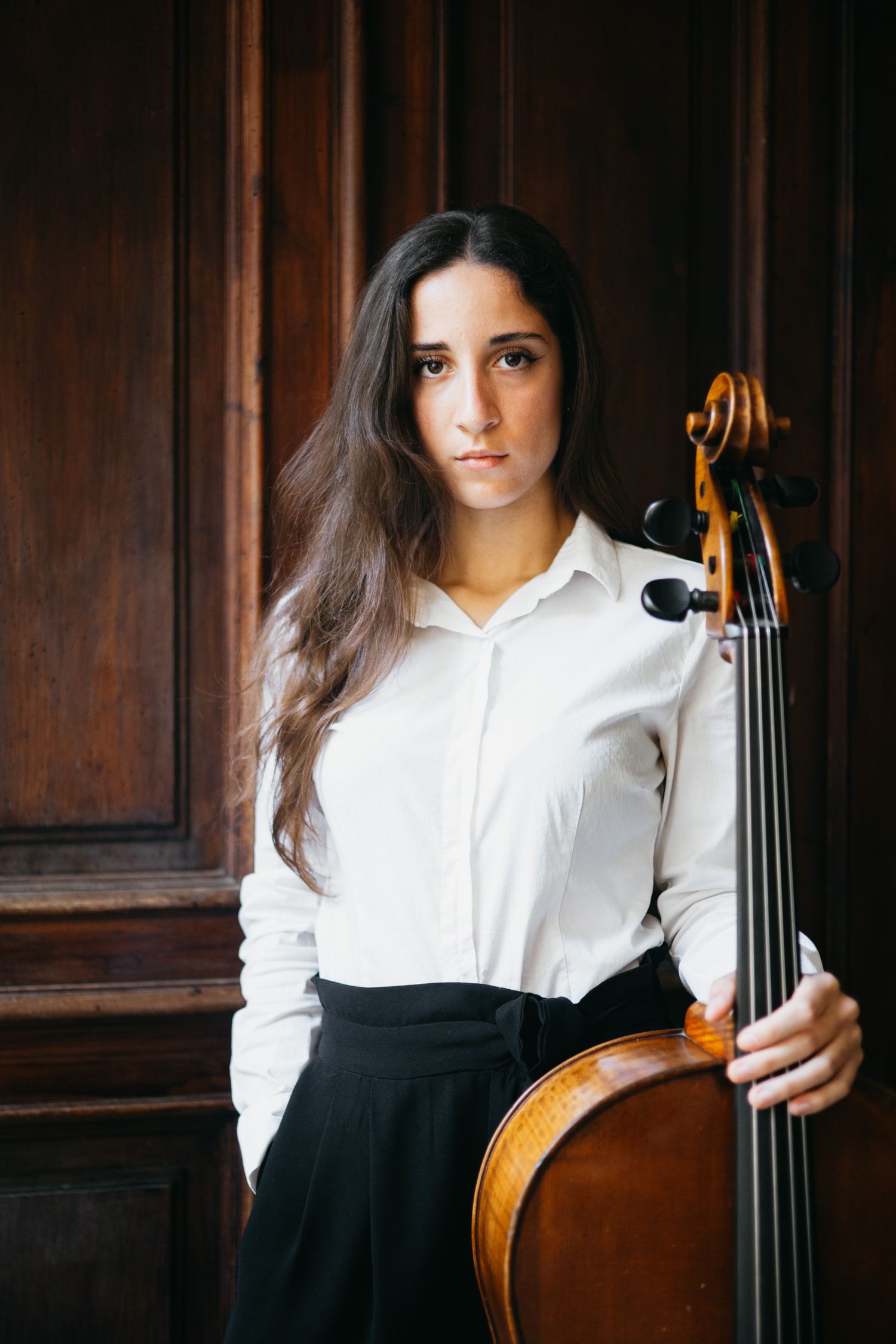 Young female cellist in white shirt standing with cello against ornate wooden paneling.