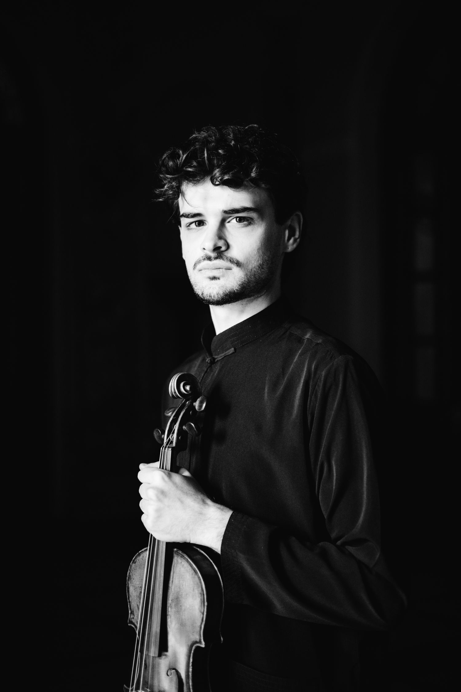 Black and white portrait of male violinist holding violin against dark background.