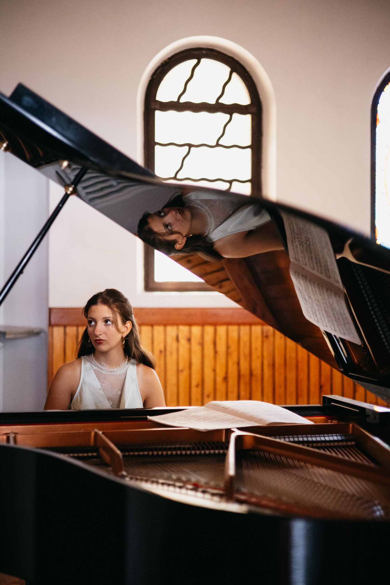 Young woman in white dress playing grand piano in a church hall with arched window in background.