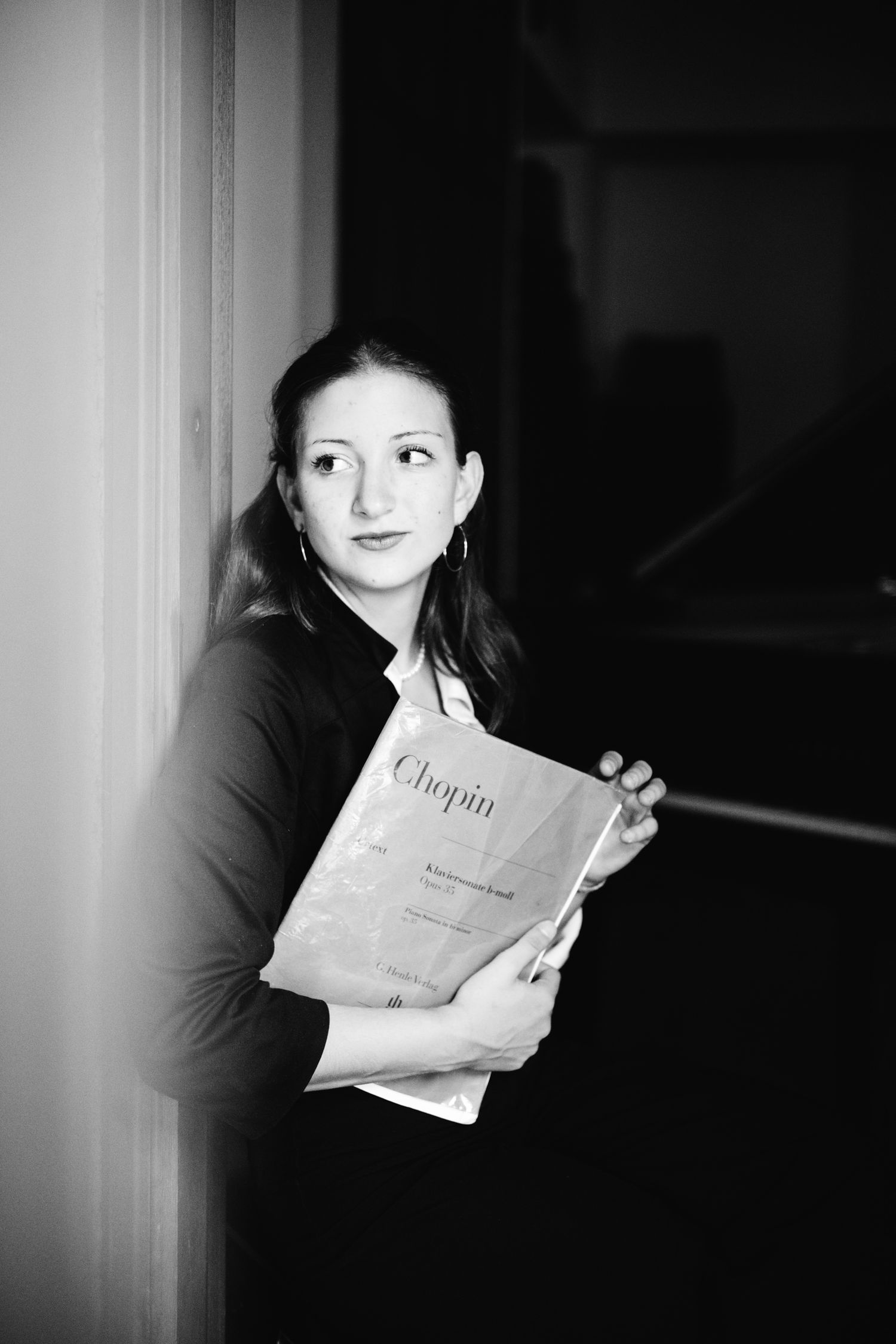 Black and white portrait of a young woman holding a Chopin sheet music book, leaning against a wall.