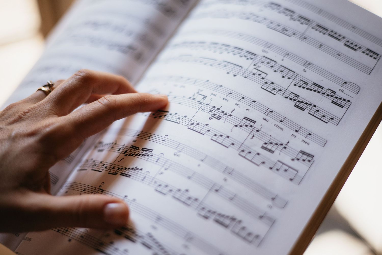 A hand with a ring follows sheet music notes in an open classical music book, reading musical notation closely.