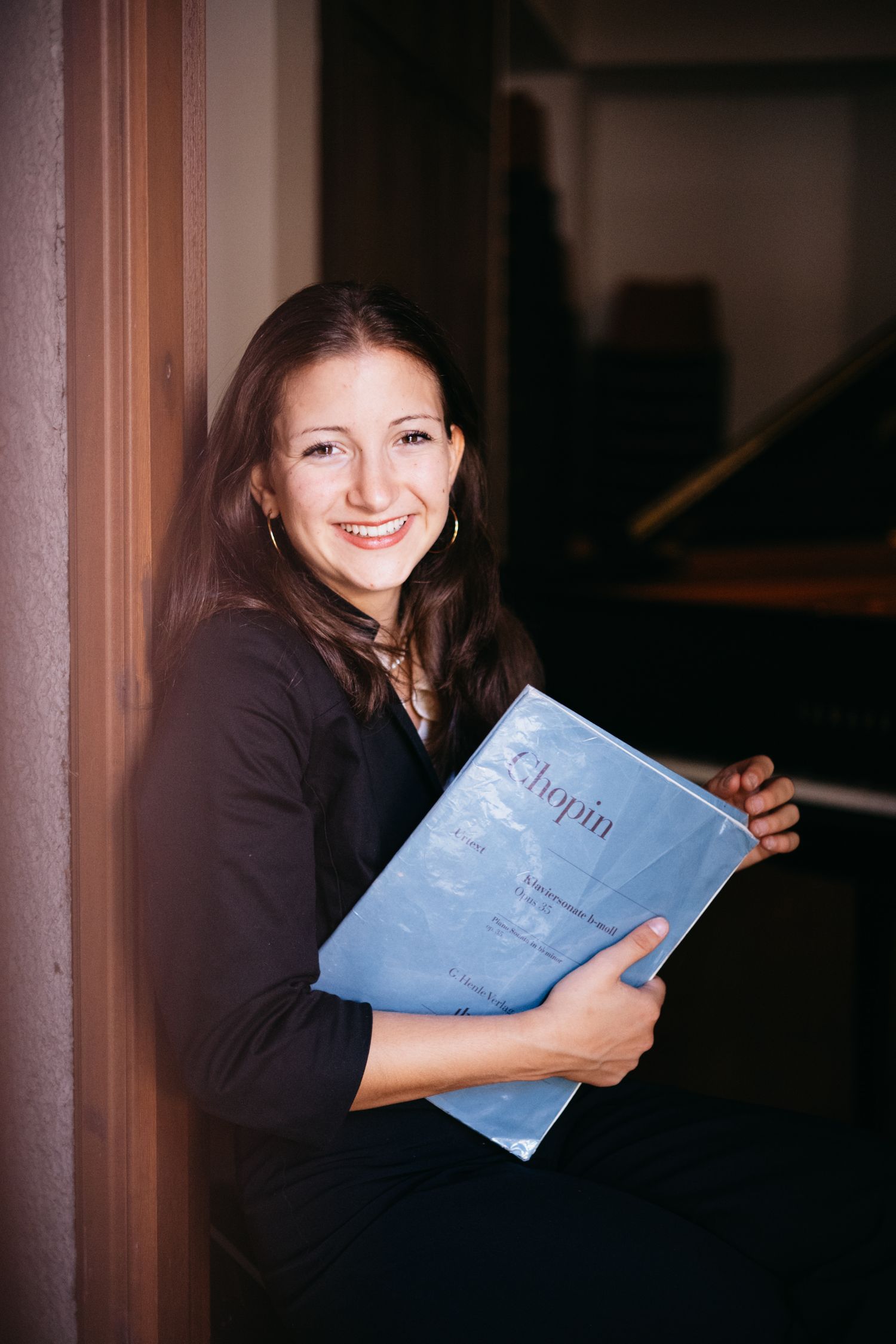 Smiling woman holding a Chopin sheet music book, seated near a piano in a dimly lit room.