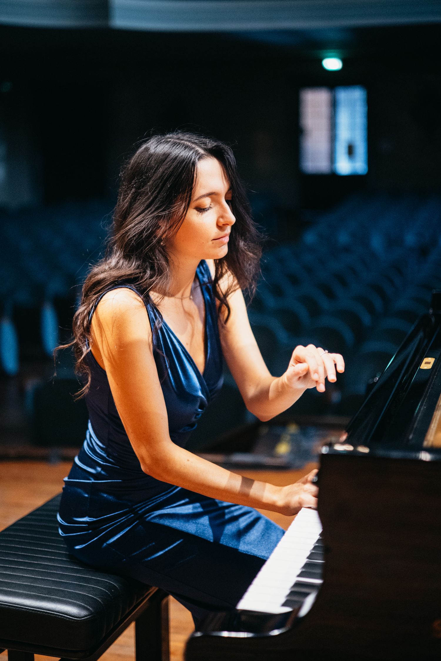 A young woman in a blue velvet dress plays a grand piano on a concert hall stage with empty seats in the background.