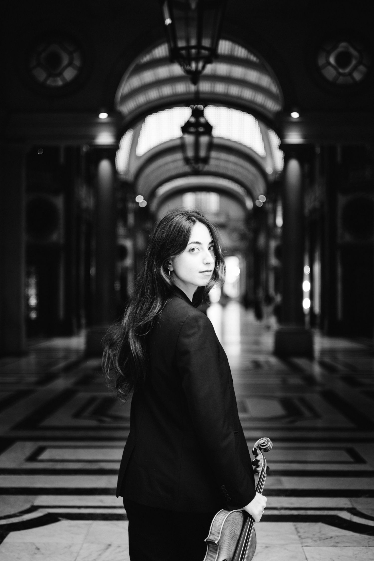 Black and white photo of a young woman holding a violin in an elegant arched hallway.