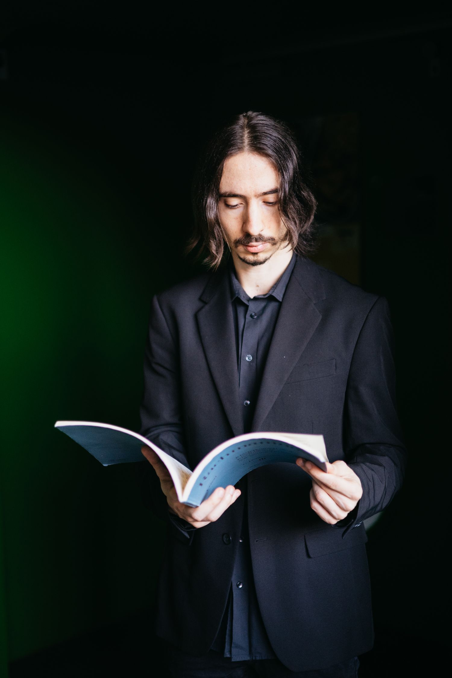 Male musician in black suit reading sheet music book against a dark green background.