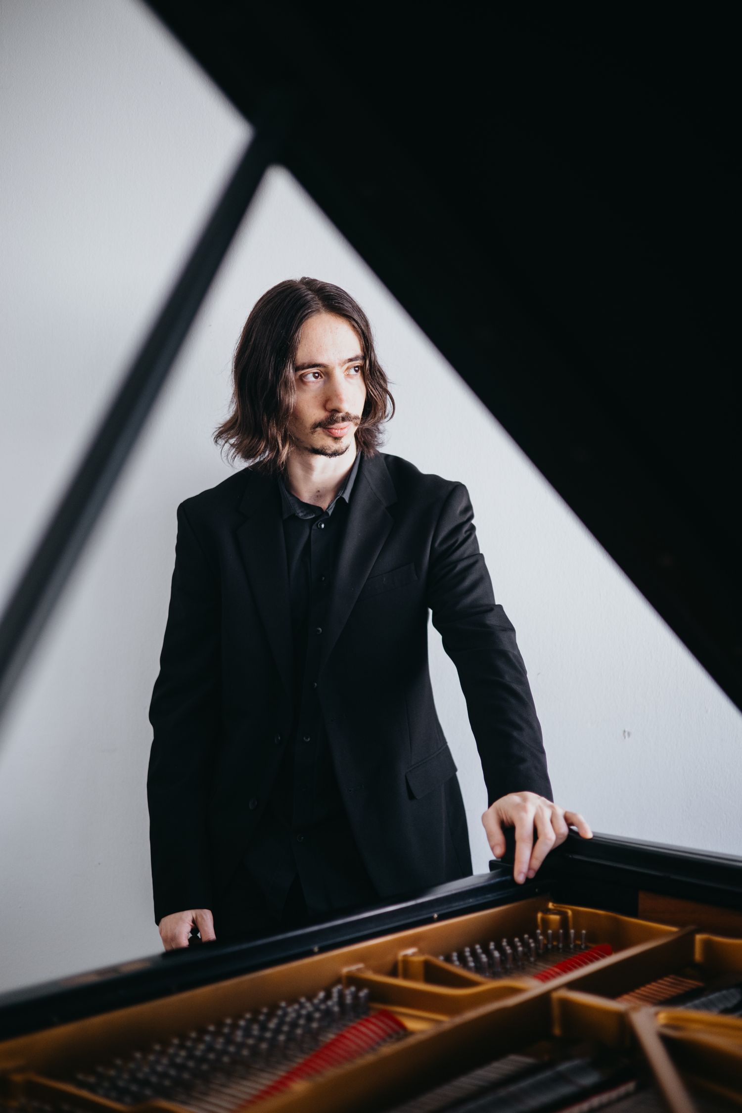 Musician in black suit standing beside an open grand piano in a dramatic lit studio.