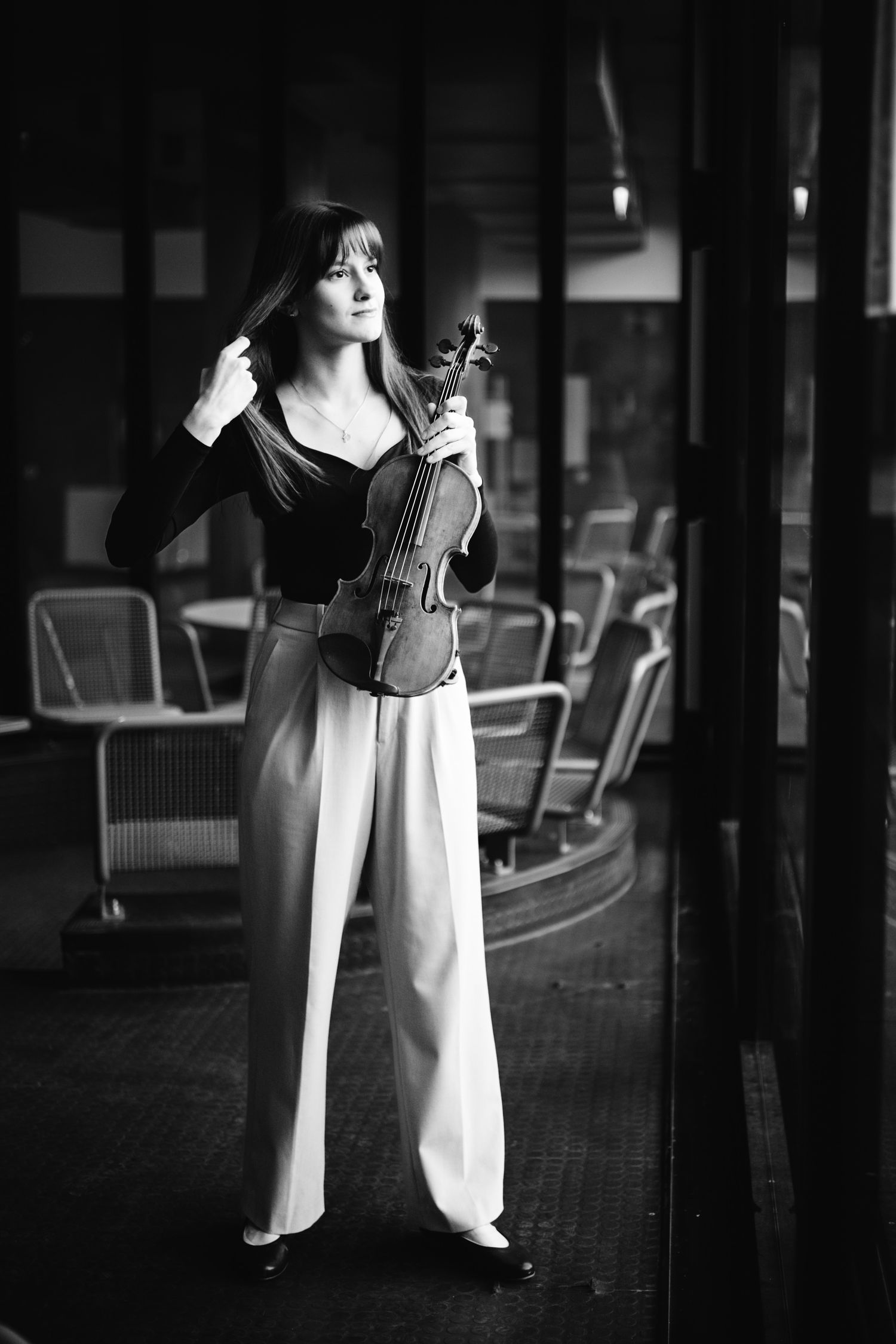 Black and white portrait of a female violinist standing in a hallway with chairs.